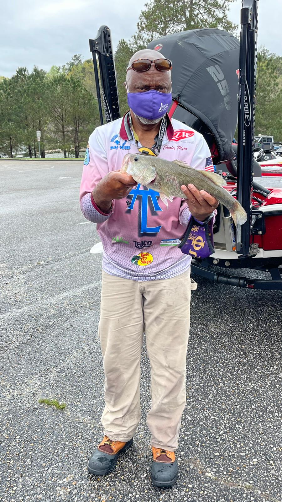 Man in purple mask and colorful shirt holds a fish near a boat in a parking lot.