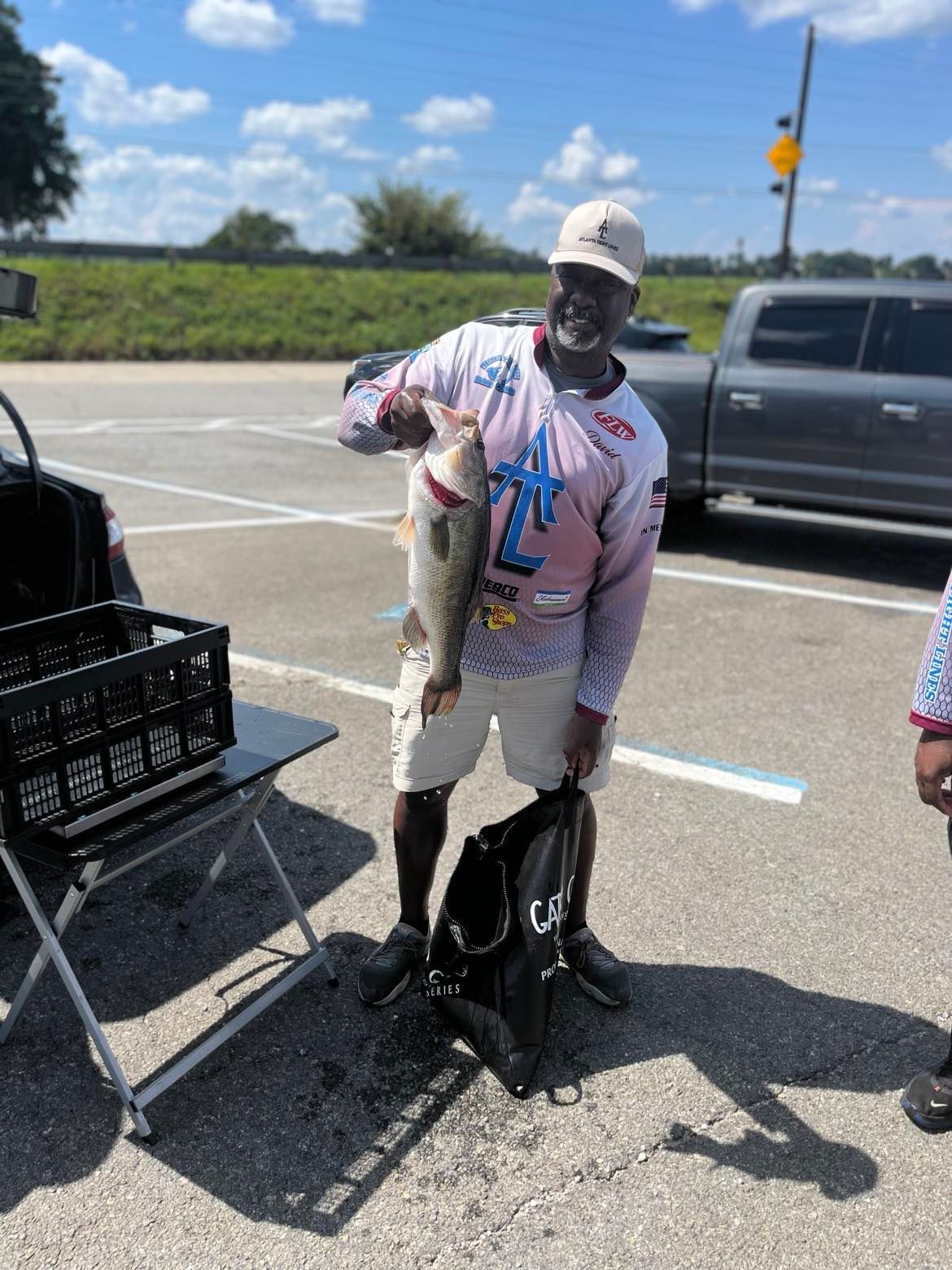 Man holding a large fish in a parking lot, wearing a long-sleeved shirt and shorts on a sunny day.