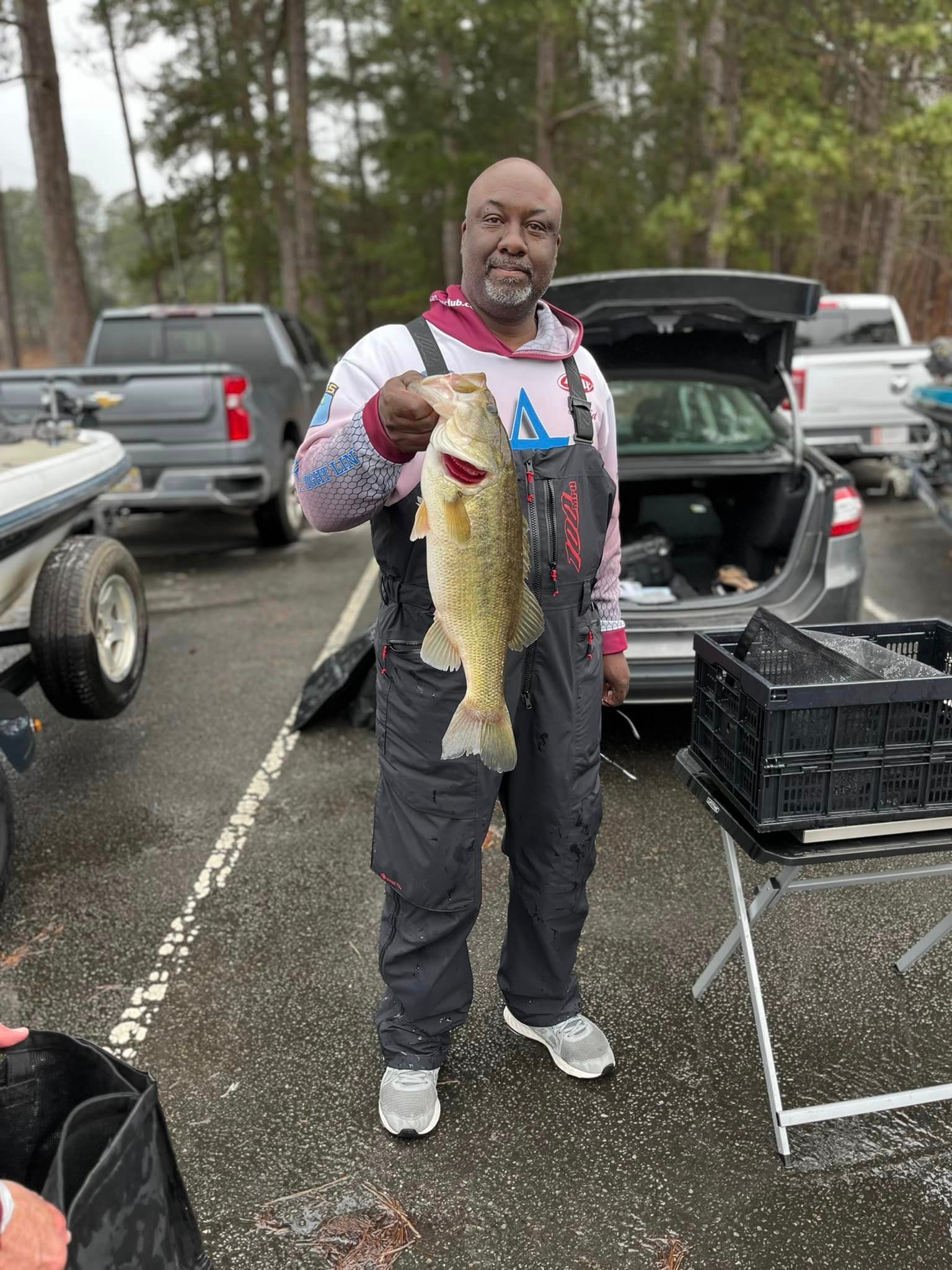 Man in fishing gear holding a large bass. Outdoors, cloudy day, near a car.