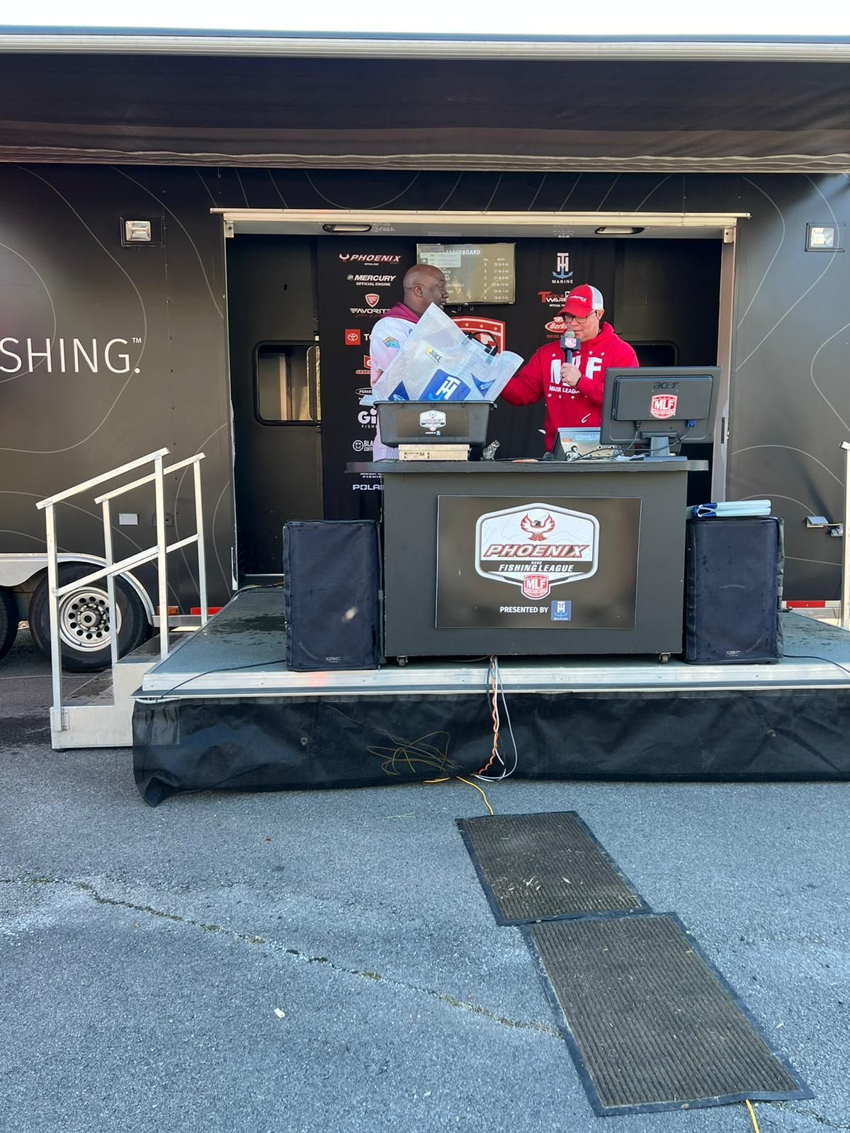 Two men in front of a trailer stage at a fishing event. One holds a trophy, the other talks.