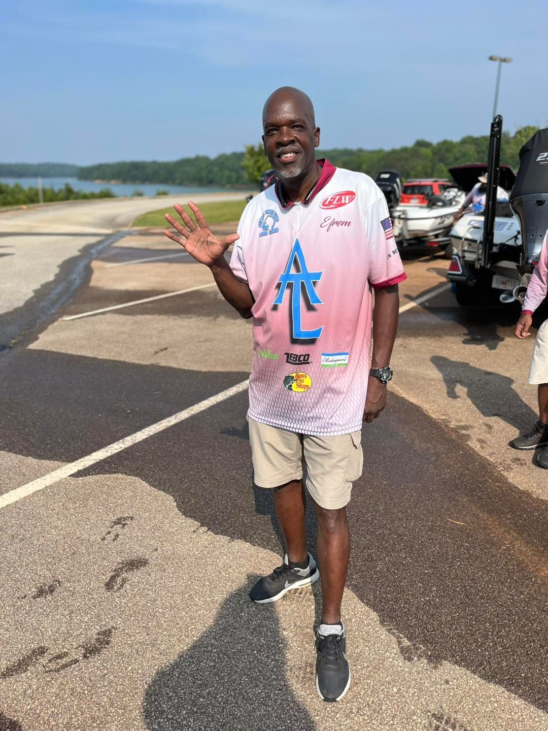 Man waves at camera, standing near lake and boats, wearing a pink fishing shirt and khaki shorts.