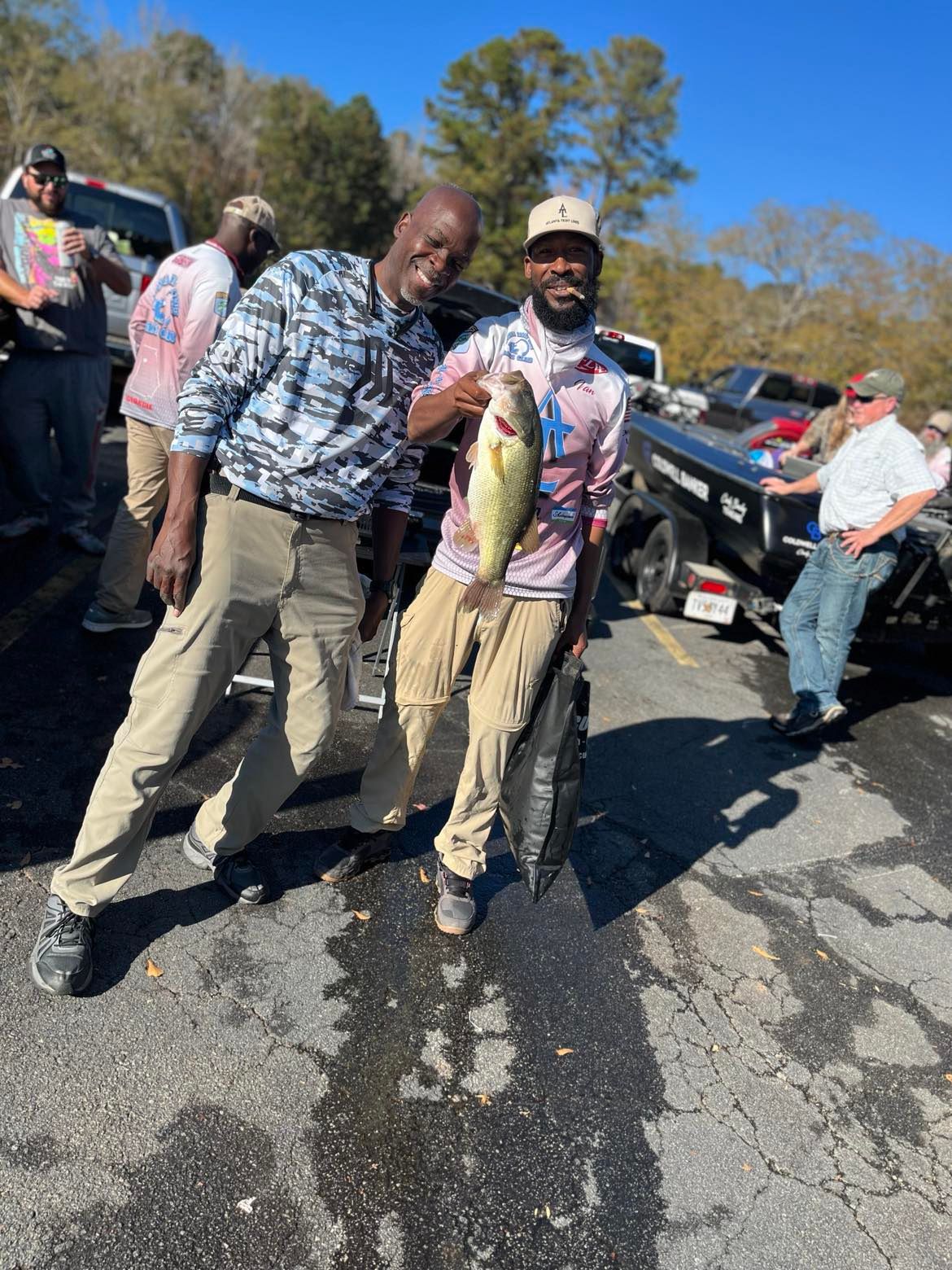 Two men smiling, holding a large fish at a boat ramp. Sunlight, concrete, trees.