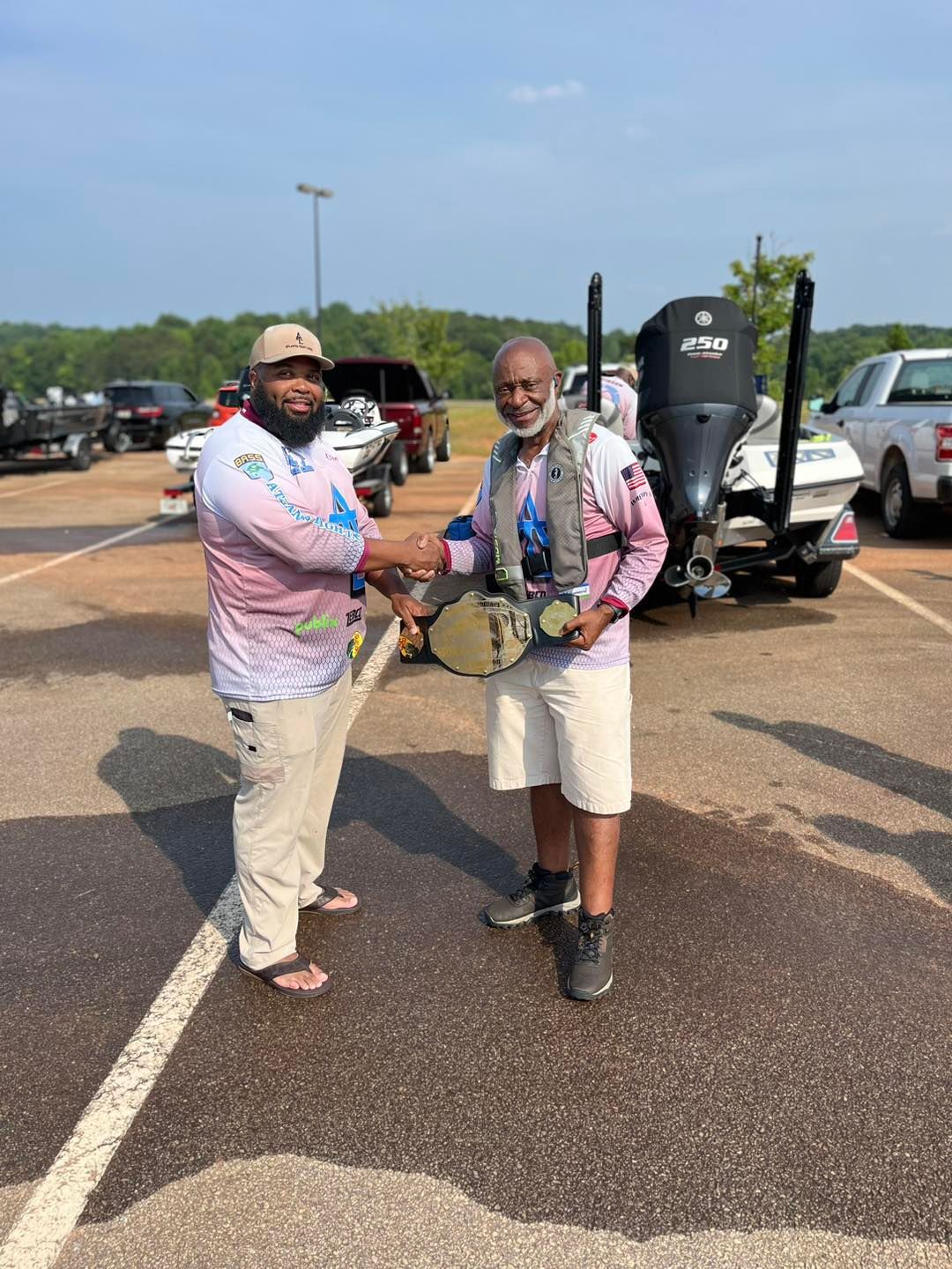 Two men shaking hands, one holding a trophy near a boat and parking lot.