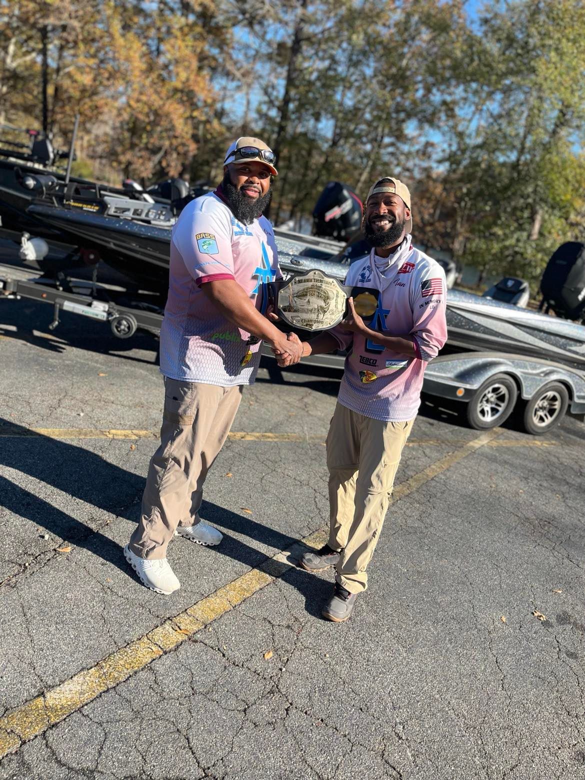 Two men shaking hands, one holding a trophy, near a boat. Outdoors on a sunny day.