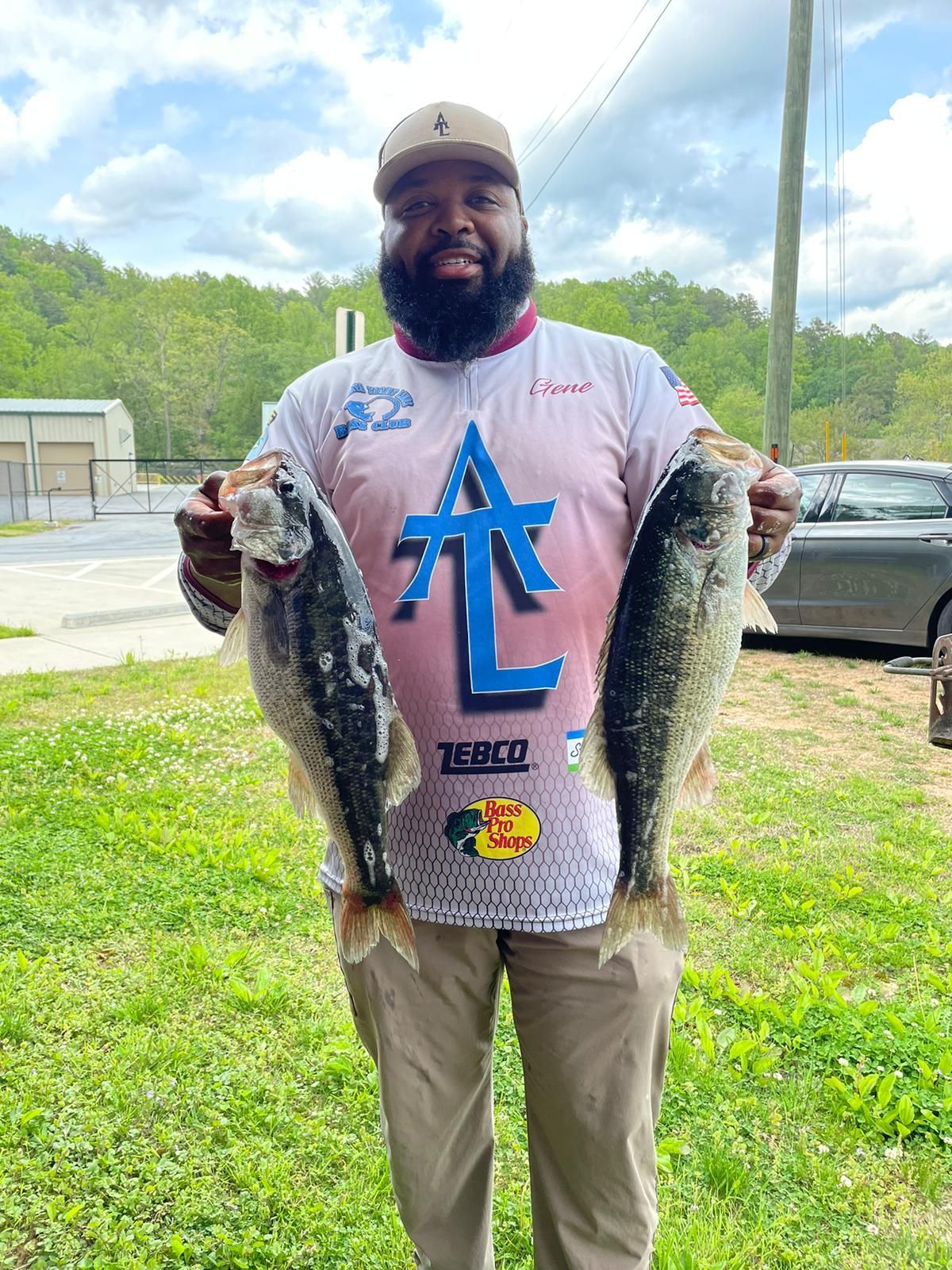 Man holding two bass, smiling, wearing a hat and fishing shirt, standing outdoors.