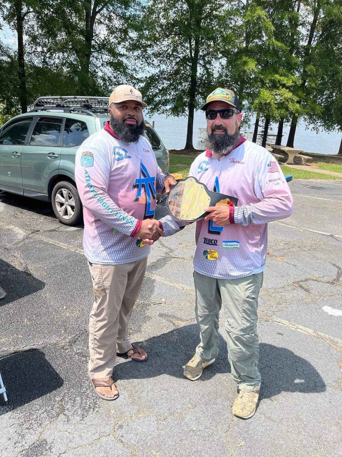 Two men shaking hands, one holding a championship belt. They wear matching pink shirts, khakis/olive pants, outdoors.