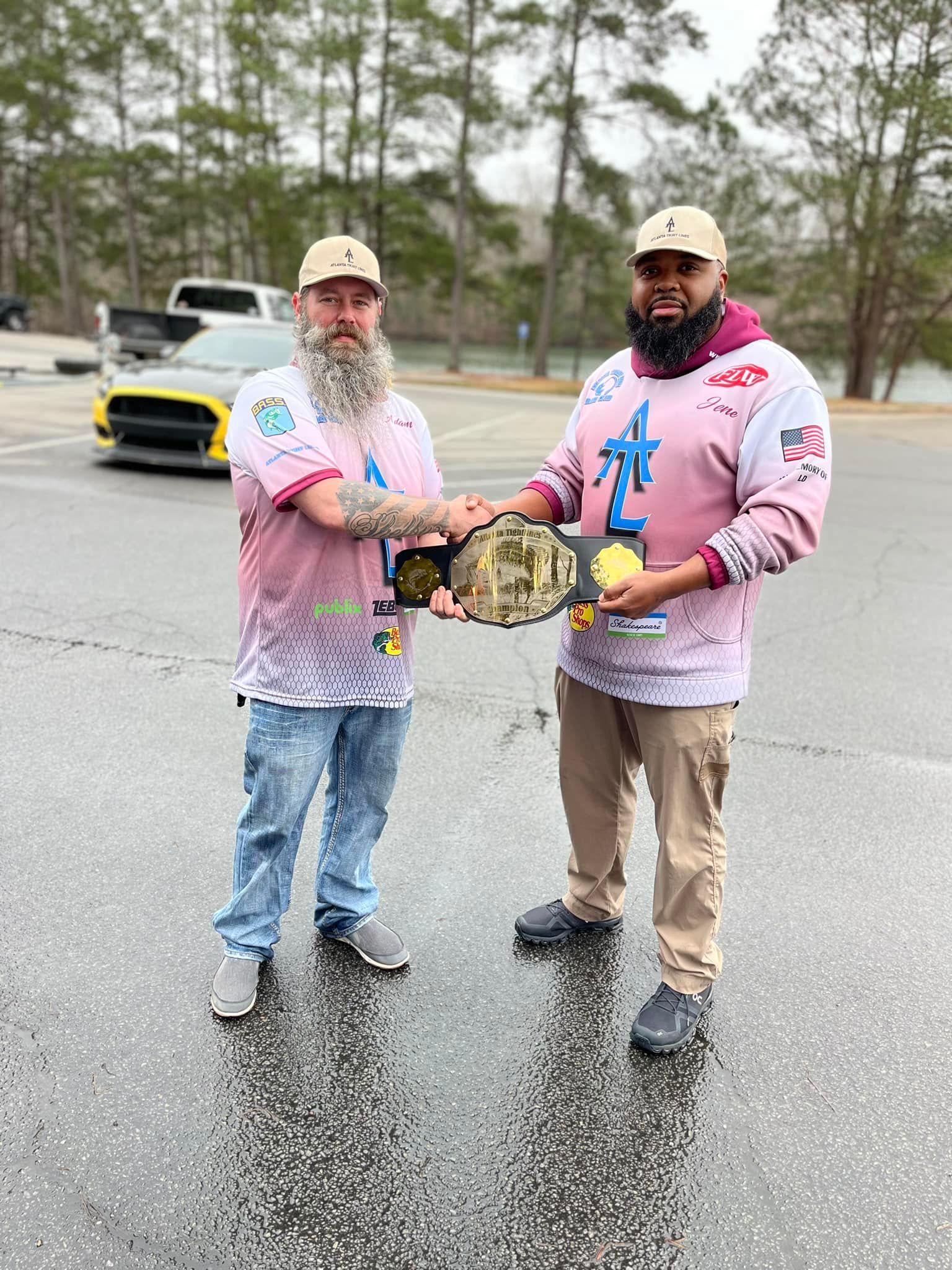 Two men holding a championship belt outside. One with a beard, the other dark-skinned. Both wear pink jerseys and hats.