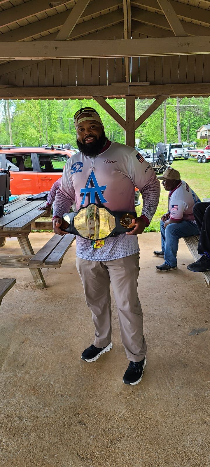 Man holding a championship belt in a pavilion, wearing a long-sleeved shirt and khakis. Other people are in the background.