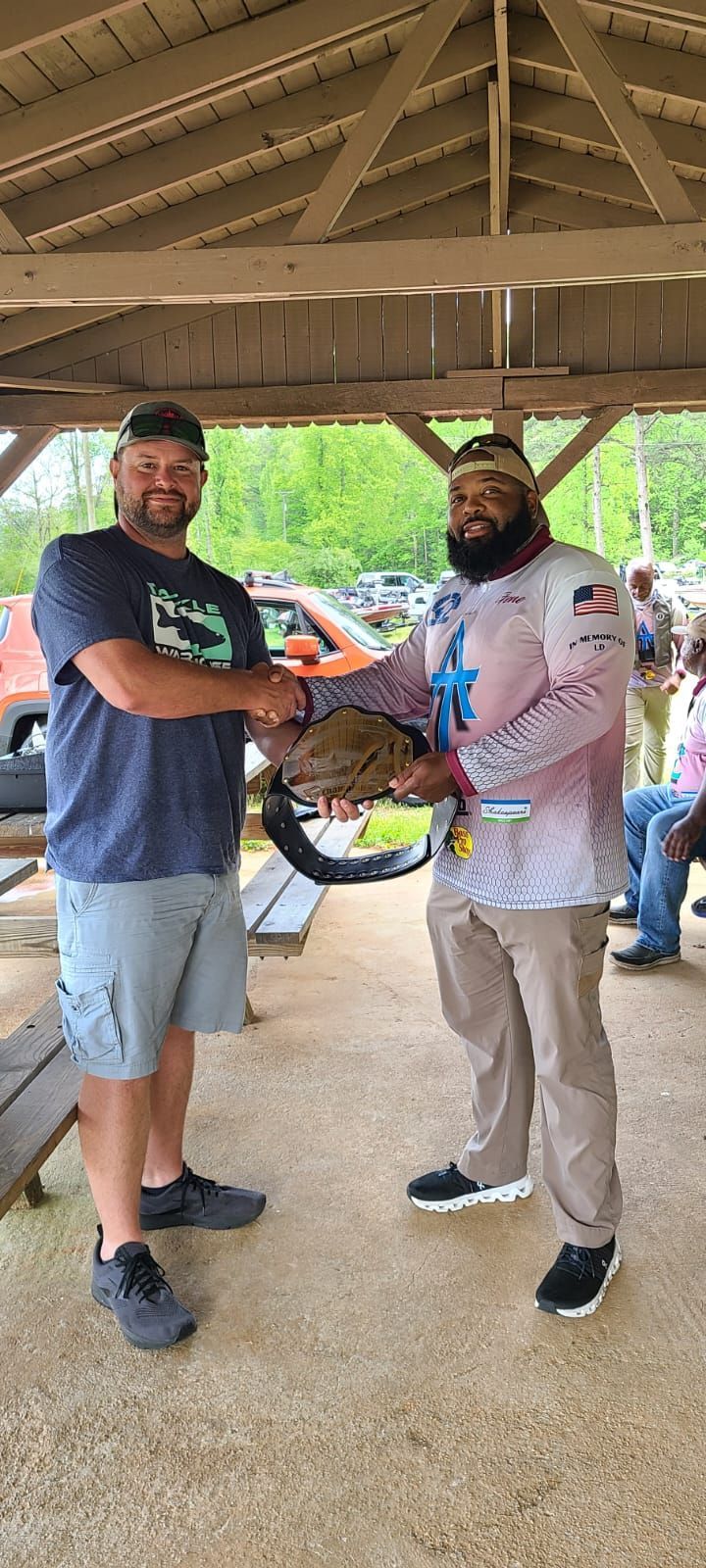 Two men shaking hands, one holding a championship belt under a wooden shelter, park setting.