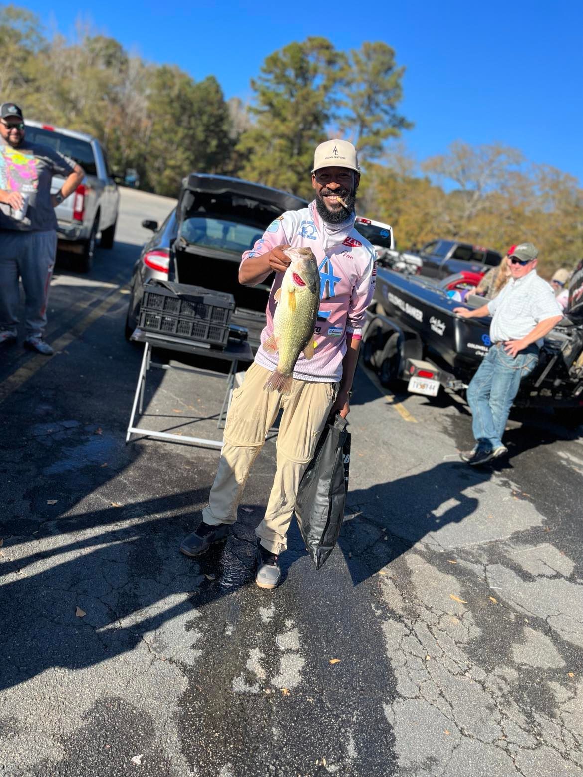 Man holding large bass, smiling, near cars at boat ramp on sunny day.