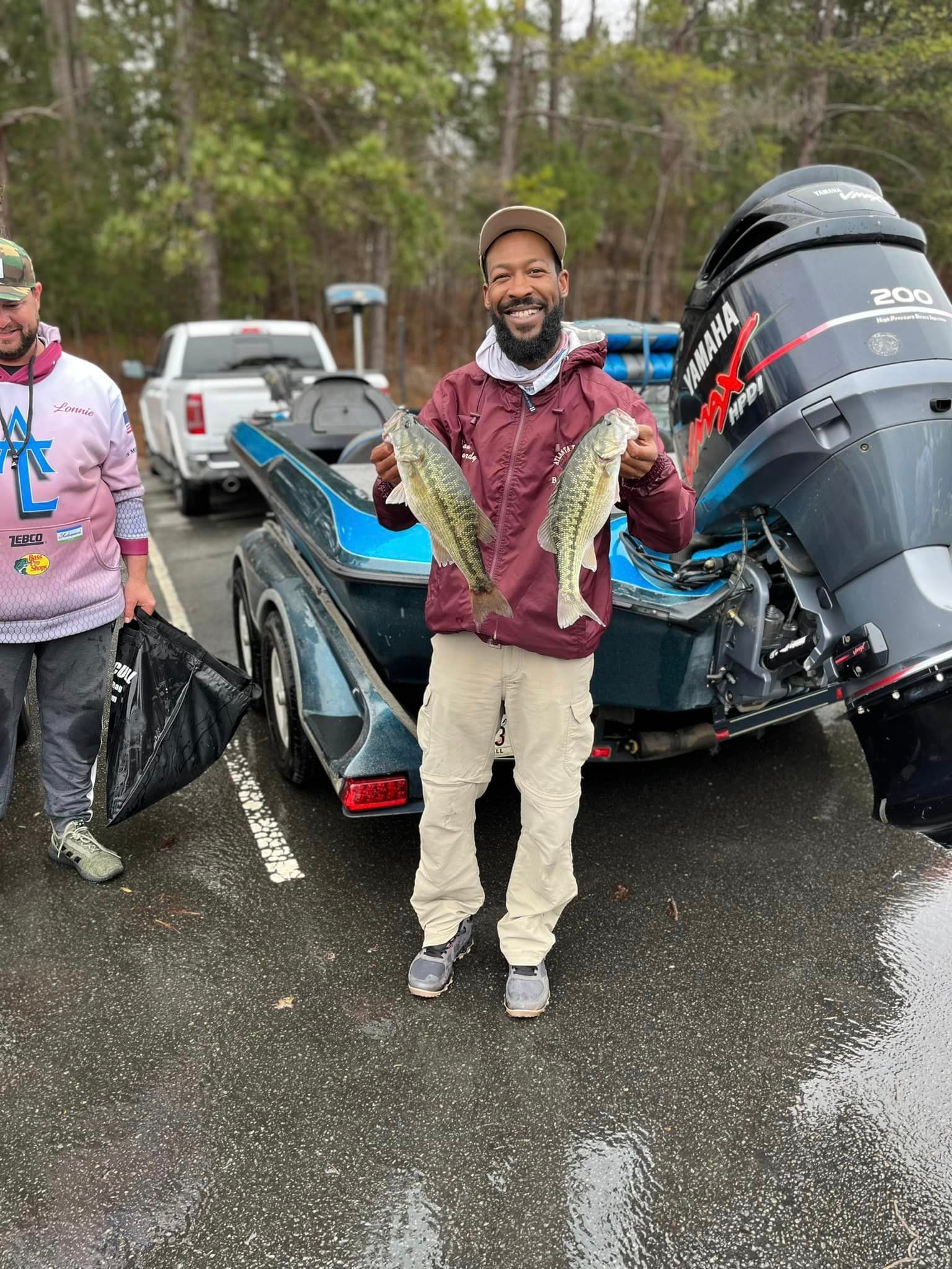 Man holding two fish near a boat, with another man holding a bag in a parking lot.