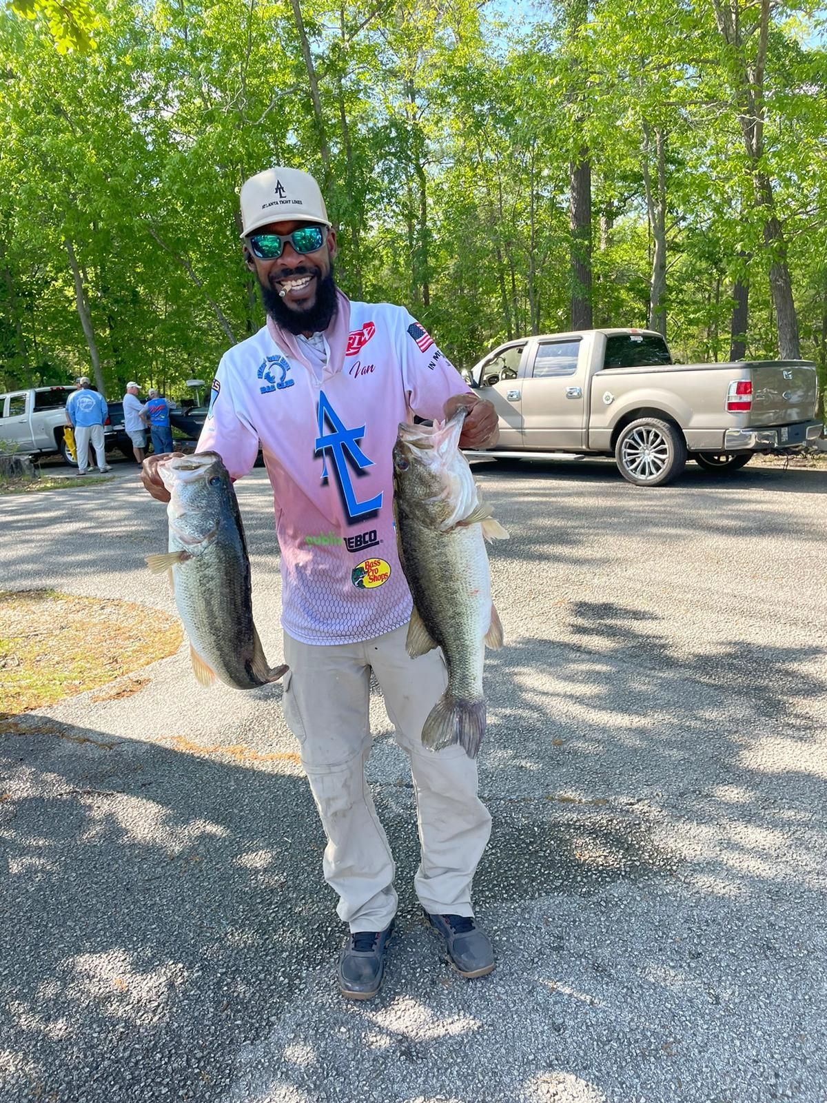 Man holding two large bass, smiling, wearing a pink shirt and hat, standing outside.