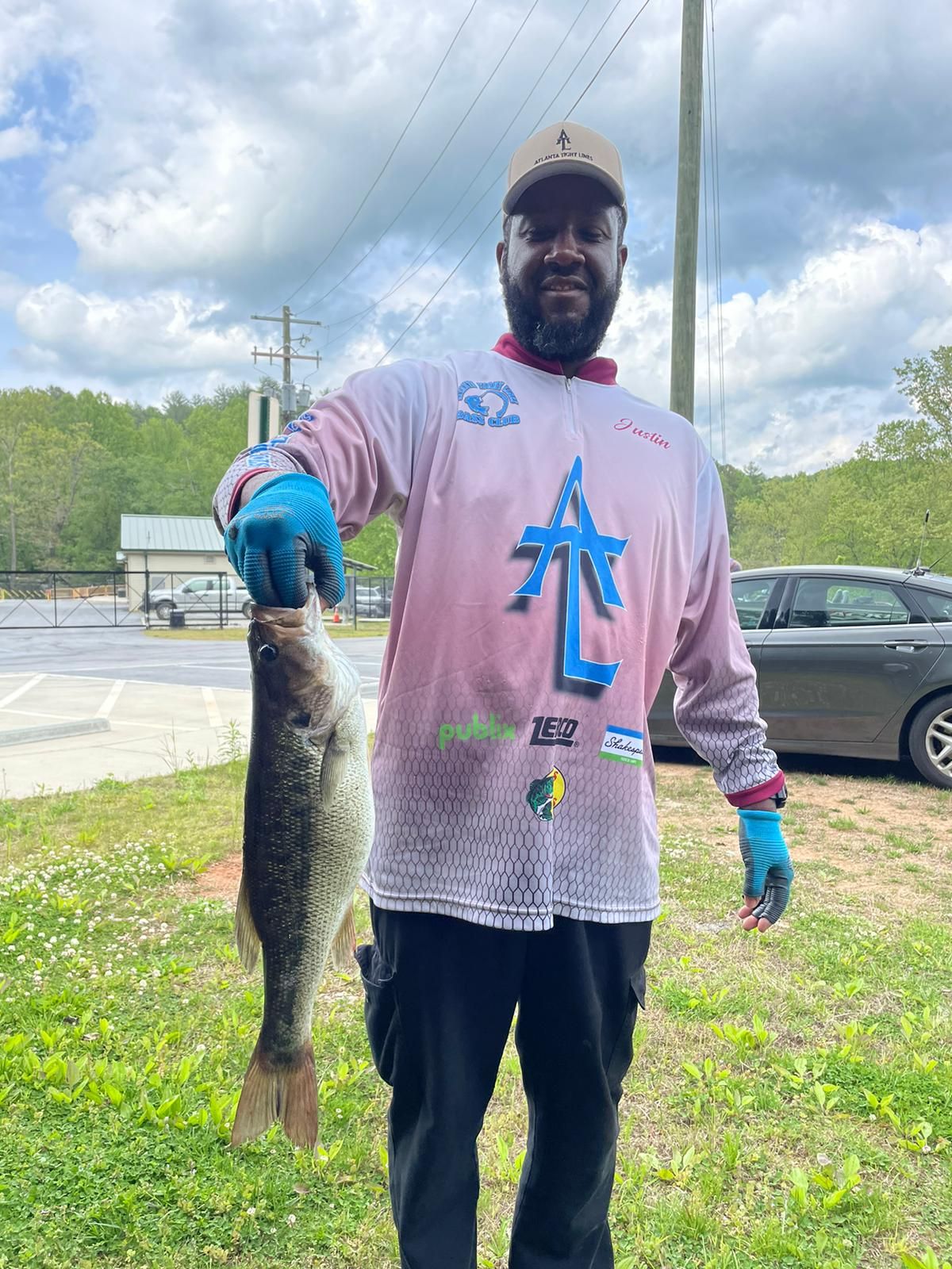 Man in pink shirt holds a large fish. Outdoors, cloudy day. He's wearing gloves, a hat, and black pants.