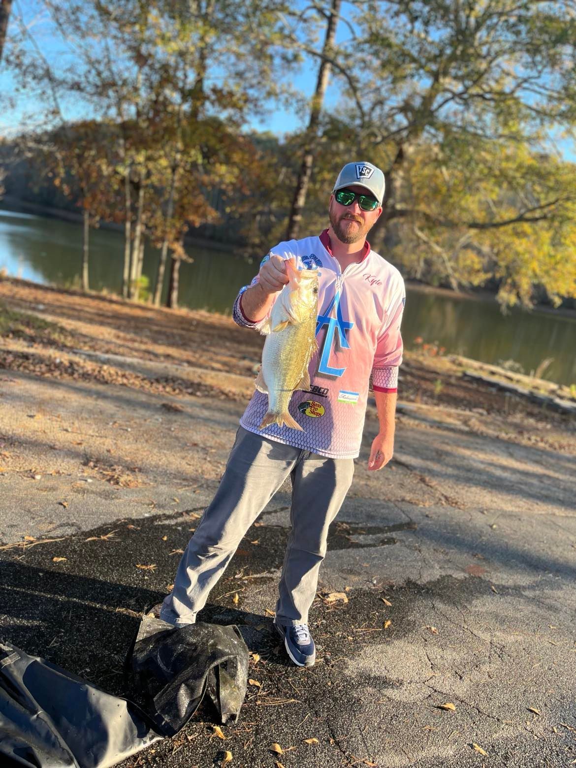Man holding large fish by a lake; wearing a hat and a pink shirt.