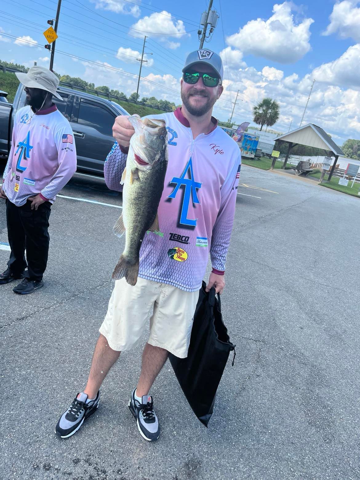 Man holding a large fish, smiling, in a parking lot. He wears a long-sleeved shirt, shorts, and hat. Another man stands nearby.