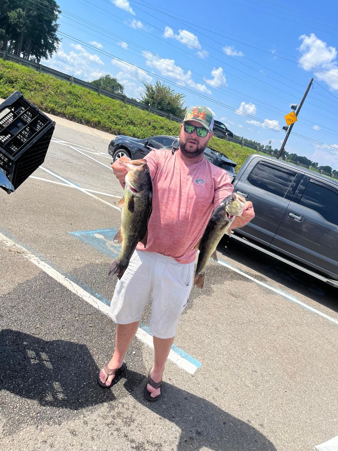Man holding two large bass fish at a parking lot, sunny day.
