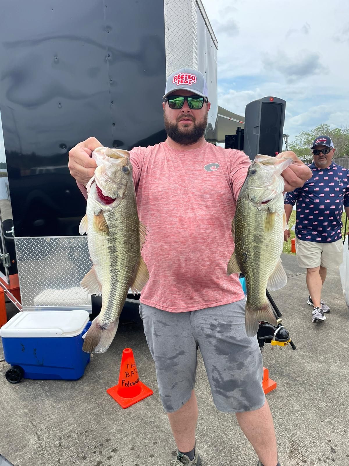 Man holding two large fish, outdoor shot near trailer. He wears cap, sunglasses, red shirt, gray shorts. Another man in background.