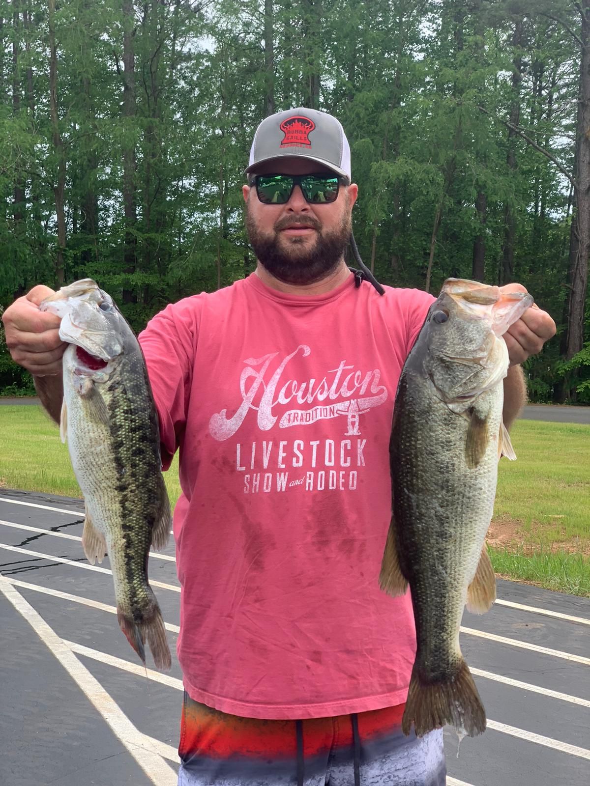 Man in red shirt holding two large bass, smiling, near a running track.