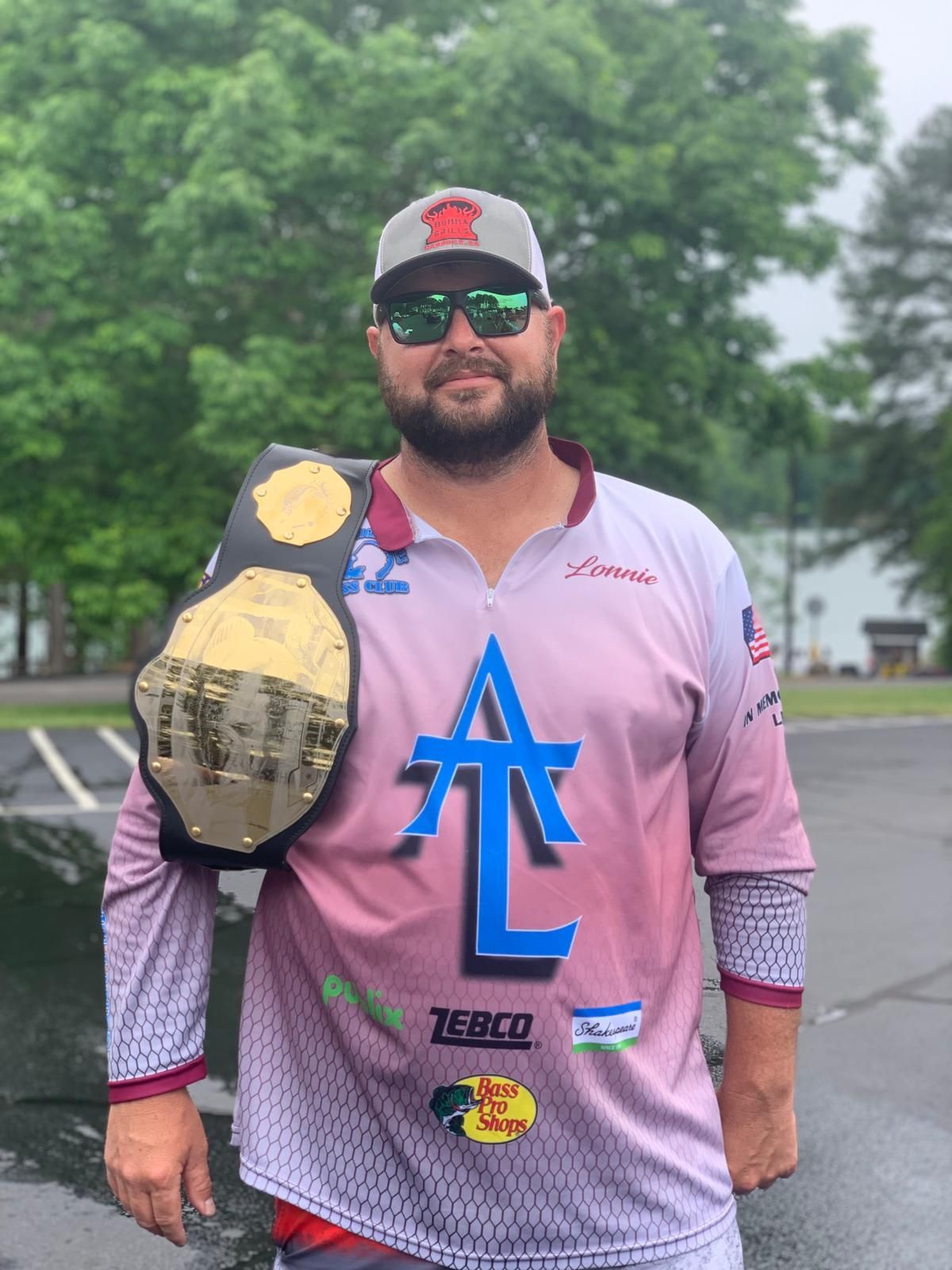 Man in fishing gear with championship belt, holding it up. Outdoor setting, overcast.