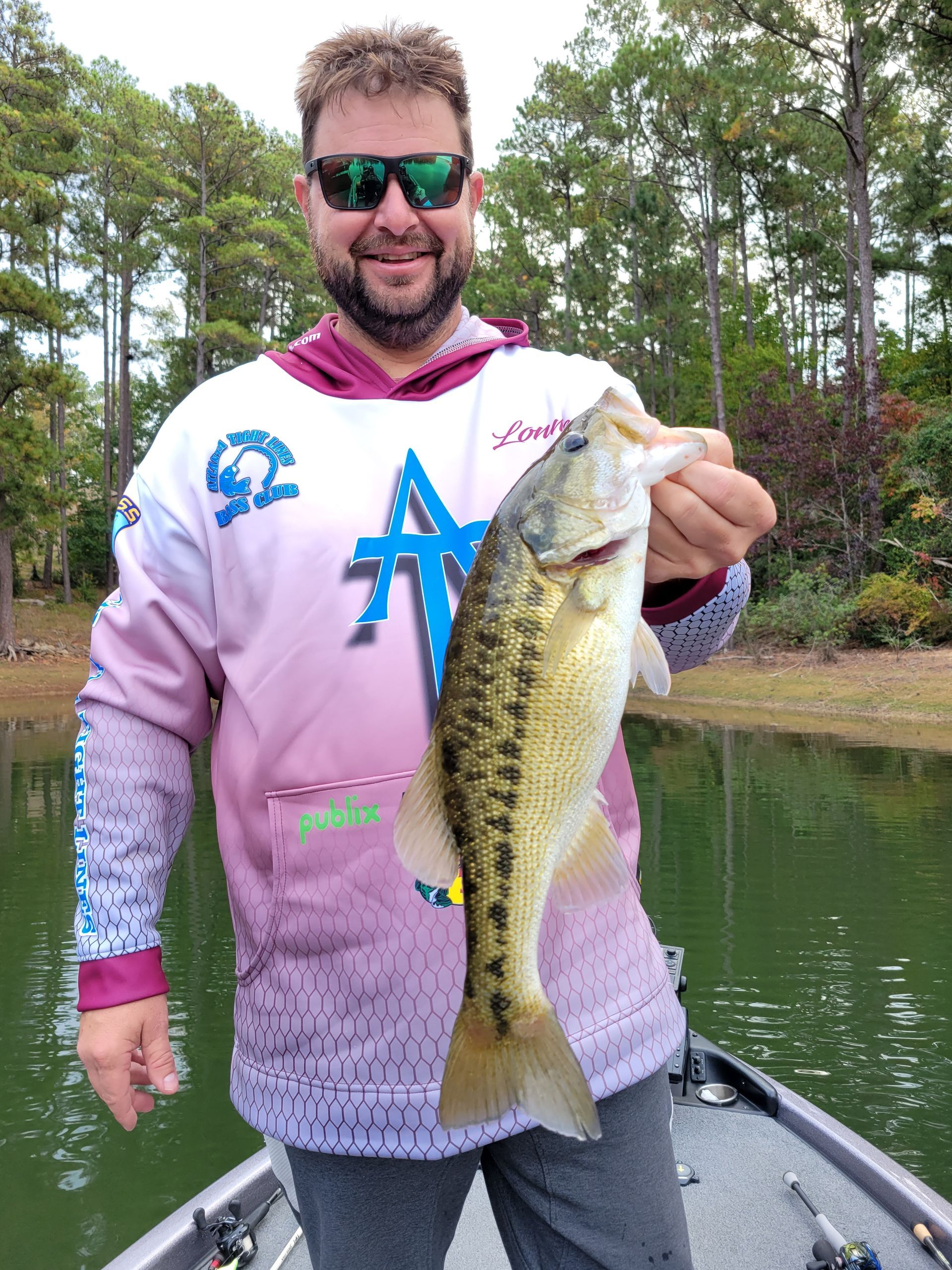 Man in sunglasses holding a largemouth bass on a boat, smiling. Lakeside with trees in the background.