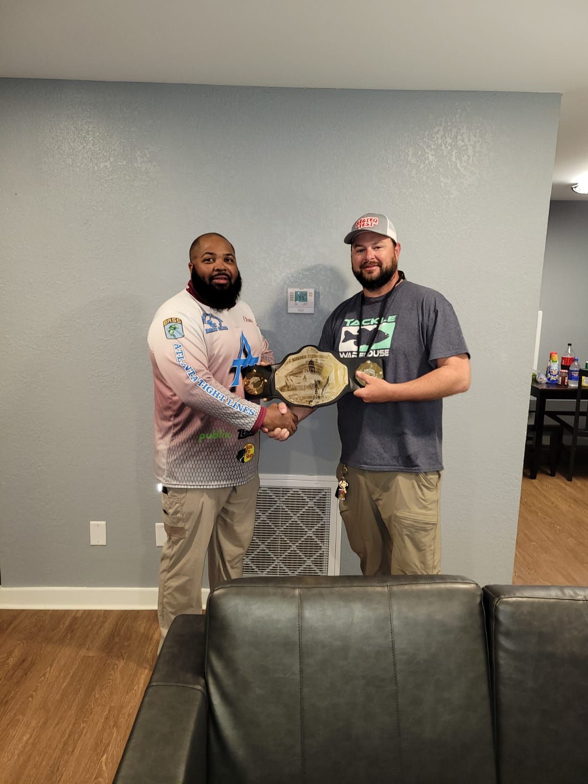 Two men shaking hands, holding a championship belt indoors. One wears a long-sleeved fishing shirt.