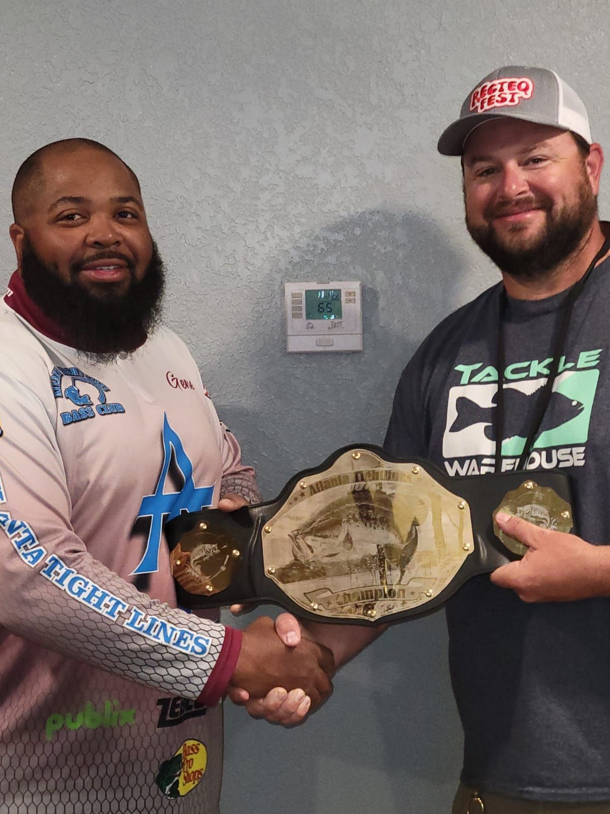 Two men shaking hands, holding a championship belt. One wears fishing apparel, the other a cap.