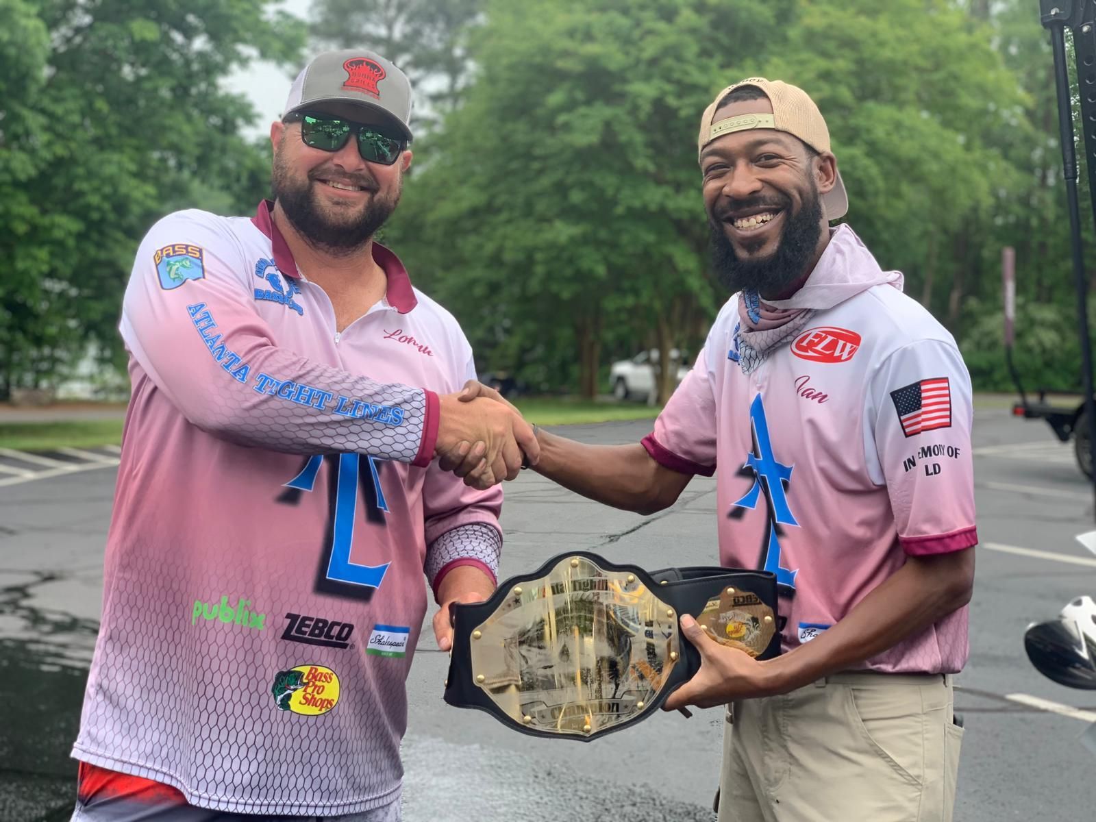 Two men shaking hands, one holding a championship belt; outdoors, both wearing fishing shirts, smiling.