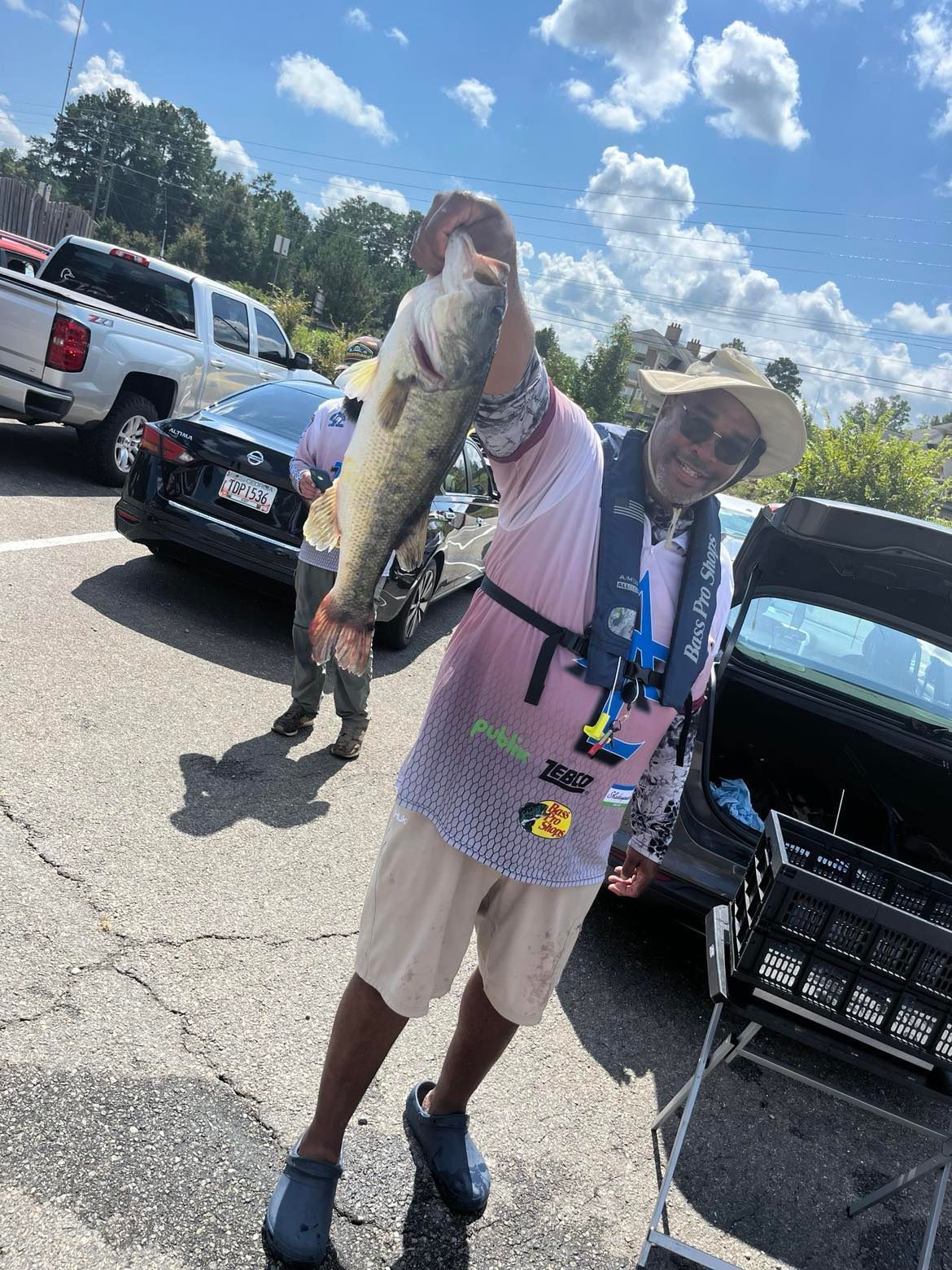 Person holding a large bass, smiling, wearing a life vest and hat. Outdoors near a car.