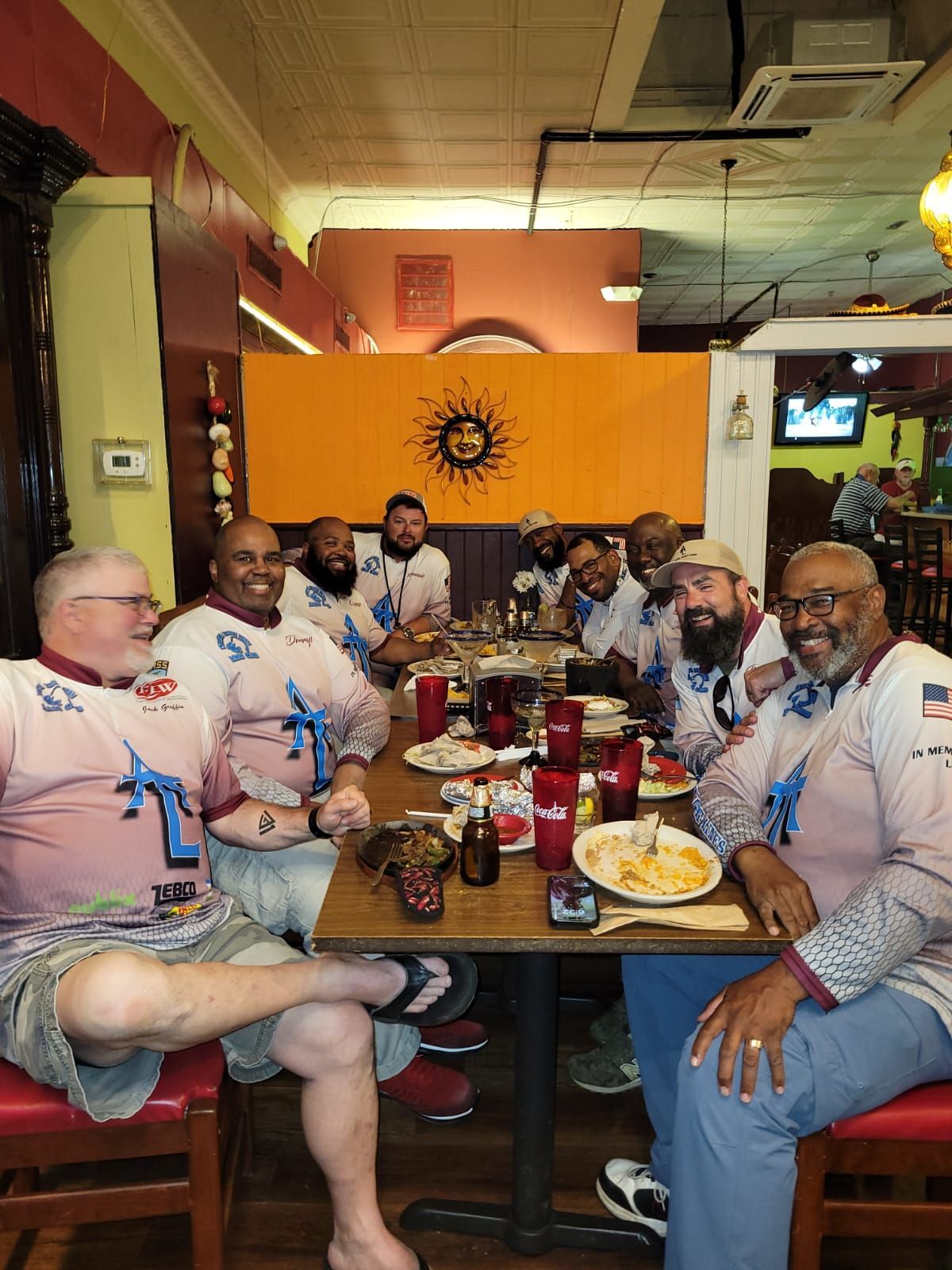 Group of men at a restaurant, wearing matching shirts, seated at a table with food and drinks.