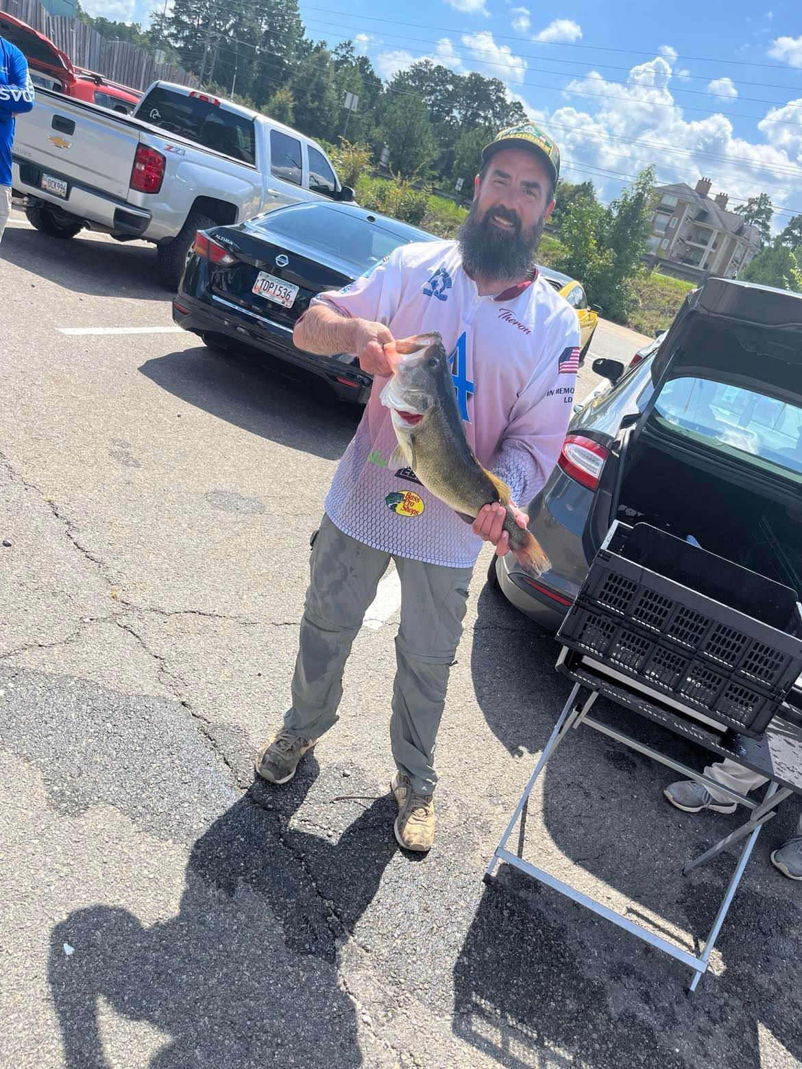 Man holding a large fish in parking lot; cars and buildings in the background; sunny day.