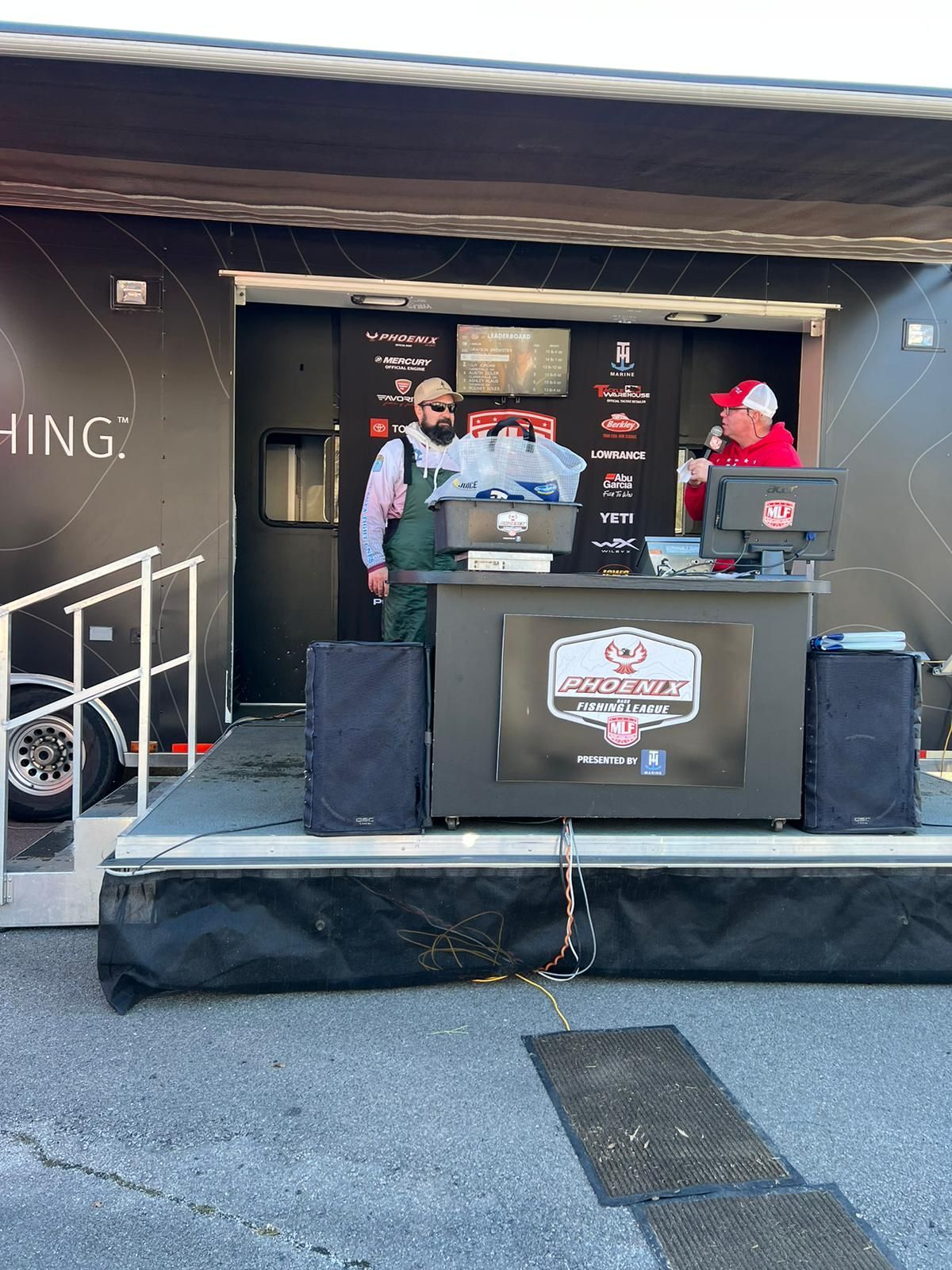 Two men at a bass fishing tournament stage. One weighs fish, the other records.