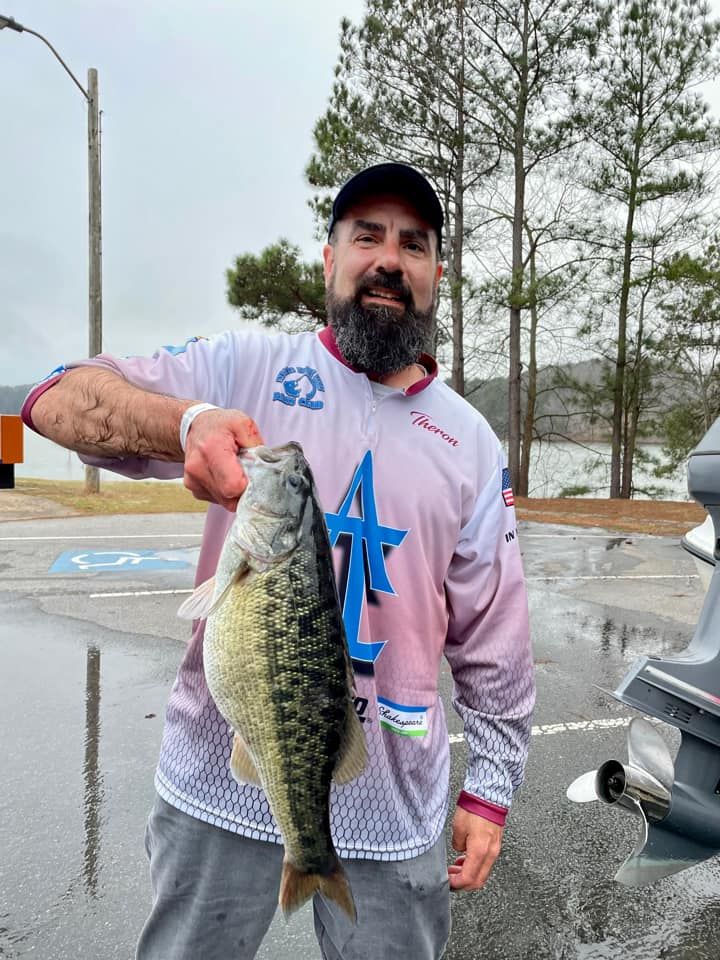 Man holding a large bass fish; outdoors, overcast day. He wears a fishing shirt, smiling.