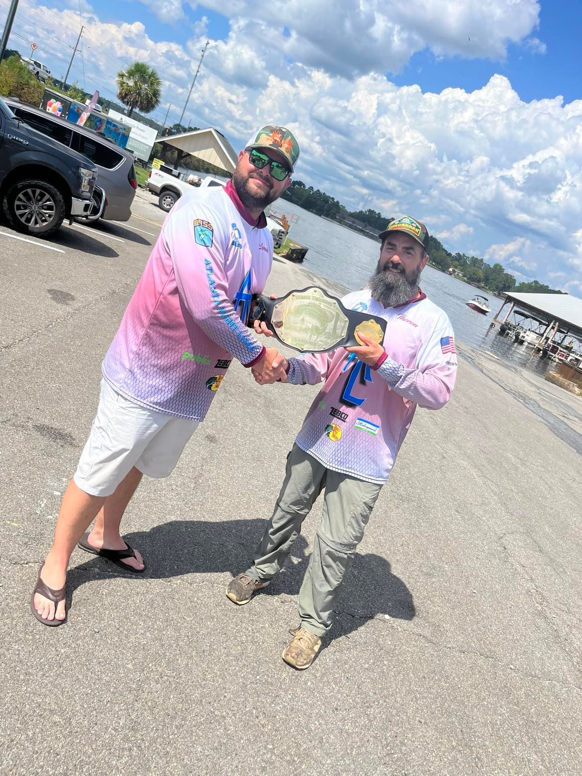 Two men shake hands, one holding a championship belt. Outdoors, near water, sunny.