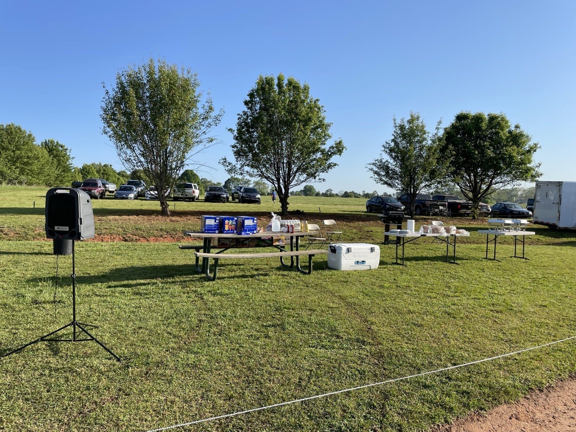 Picnic setup in a grassy field with tables, cooler, speaker, and trees; cars parked in the background under a blue sky.