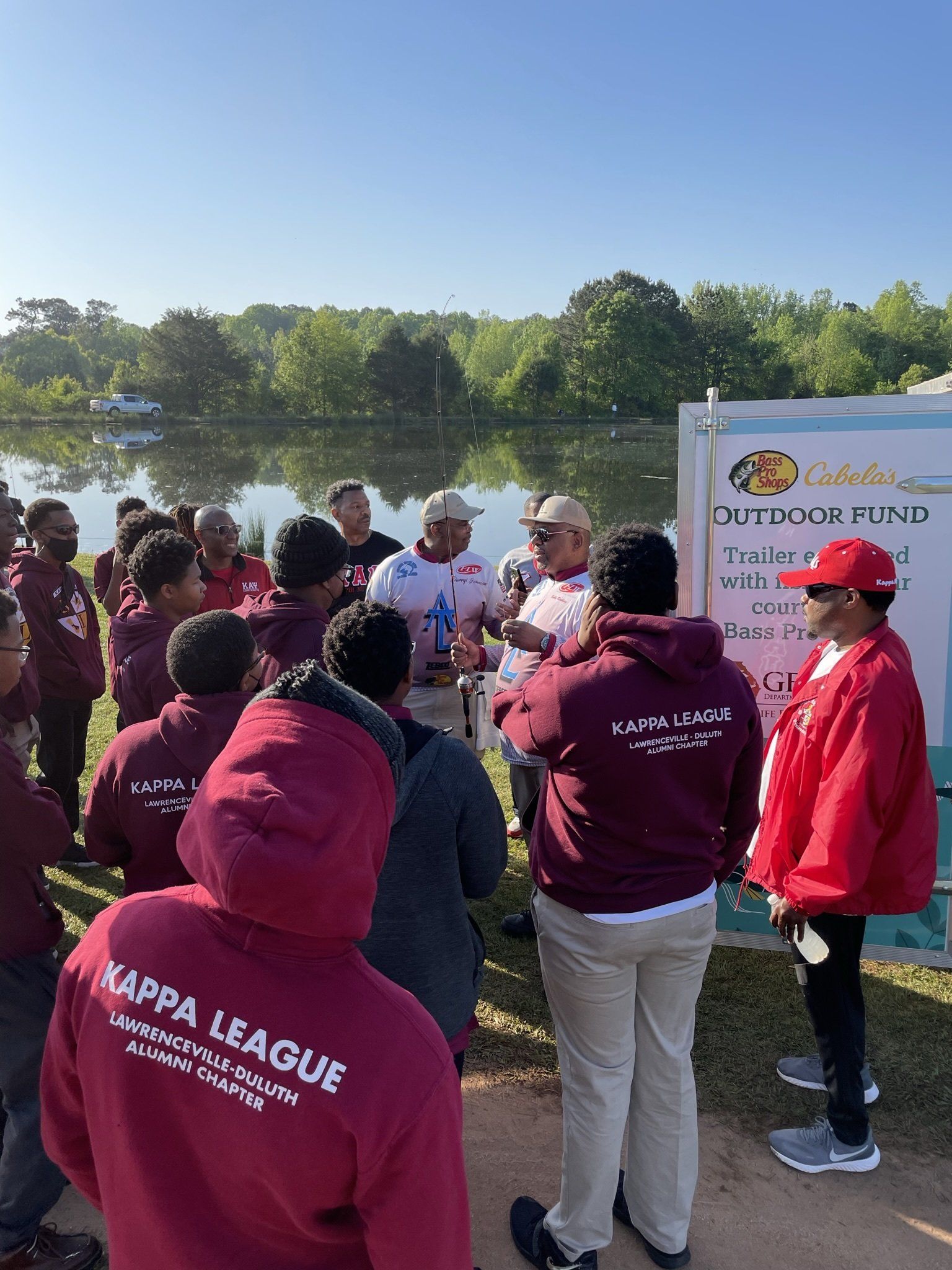 Group of people in burgundy hoodies and red jackets gather near a lake for an outdoor event.