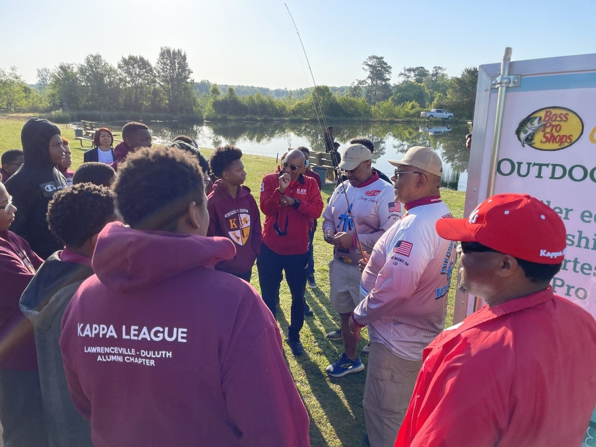 Group of people at lakeside, learning fishing. Some wear red caps/jackets, one holds a fishing rod.
