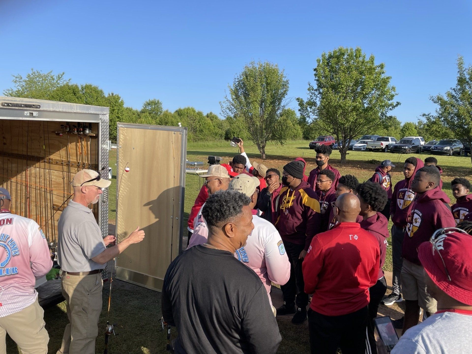 A group of people in a park listen to a man speaking near a trailer. Many wear red and gold.