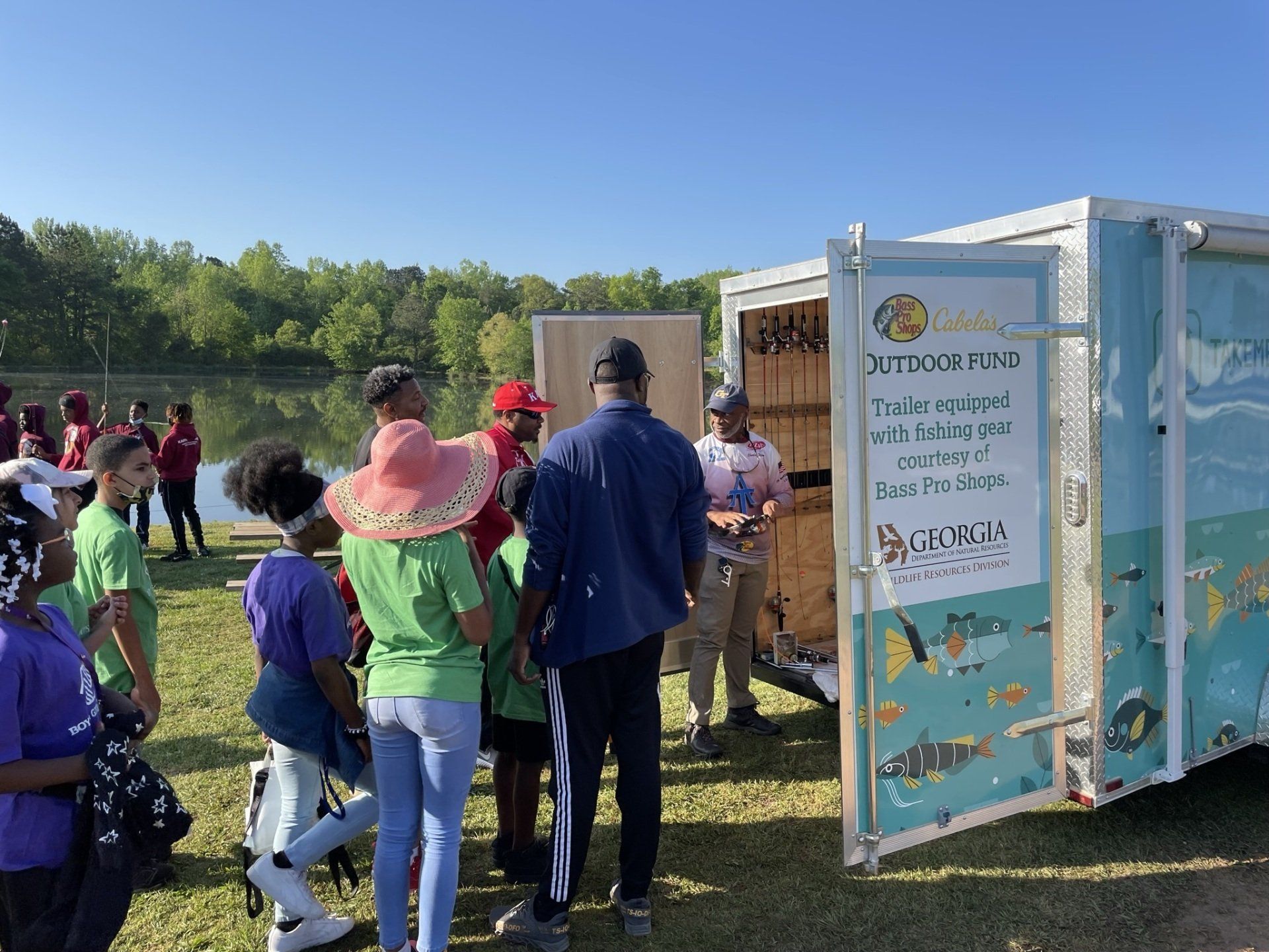 Group of kids listen to a speaker outside a Bass Pro Shops trailer by a lake.