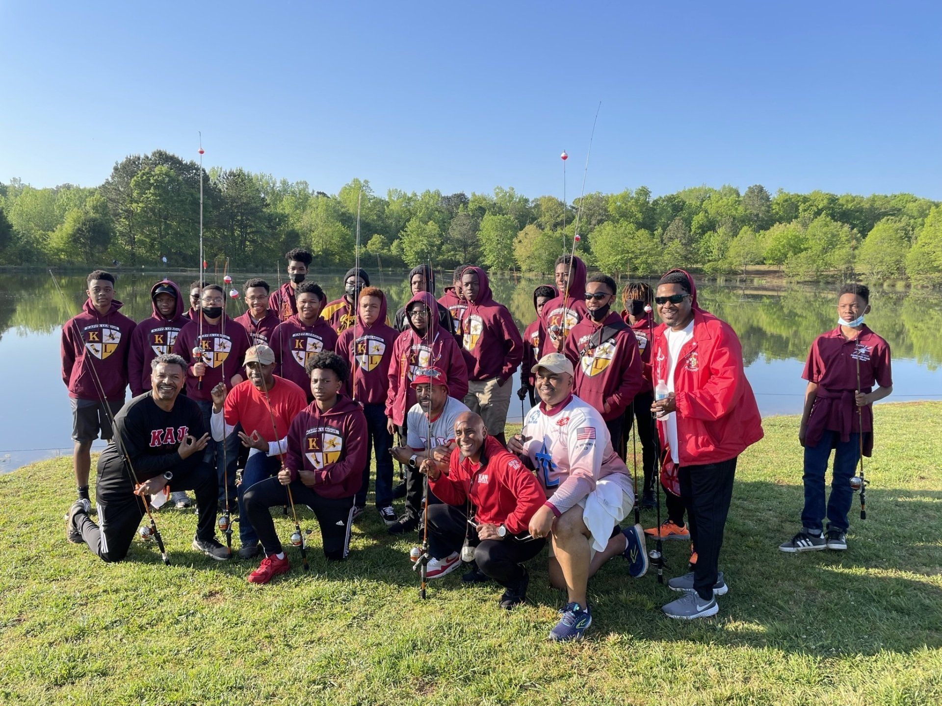 Group of people wearing maroon hoodies and red jackets pose by a lake, holding fishing rods.