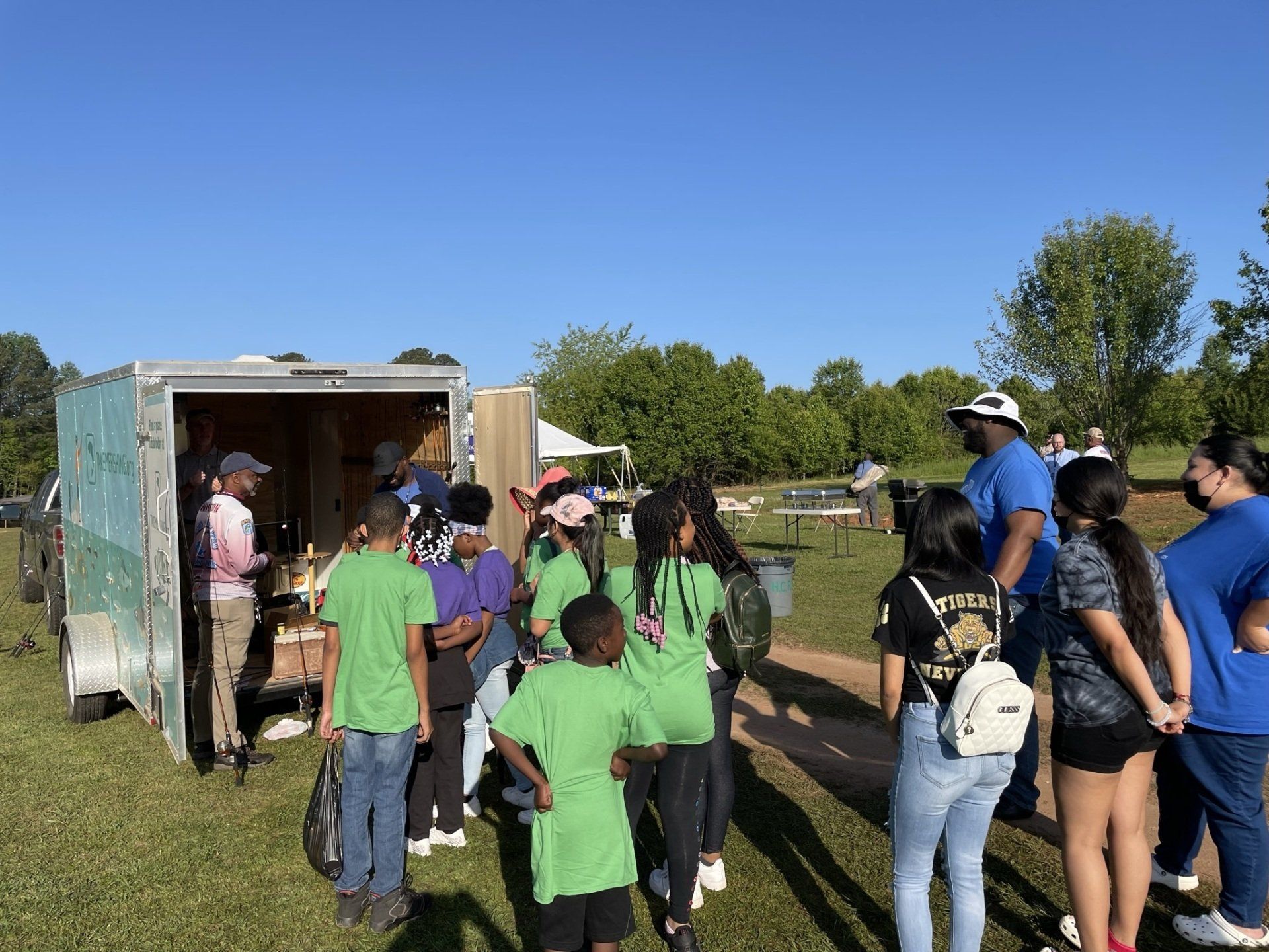 Group of people gathered outdoors near a trailer, some wearing green or blue shirts, on a sunny day.