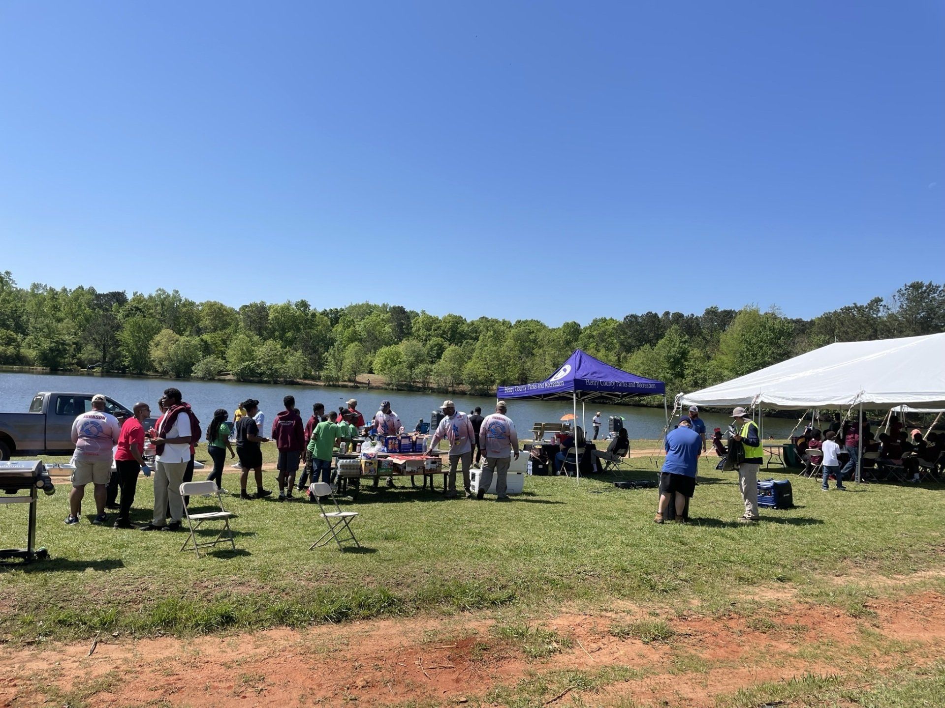 People gathered at a lakeside event. White tent and purple canopy offer shade. Blue sky, green trees.