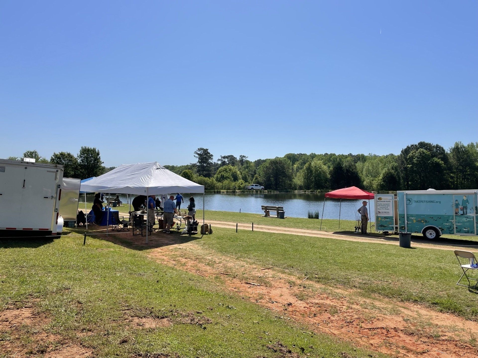 Event on a grassy field by a lake; tents, trailers, people, clear sky.