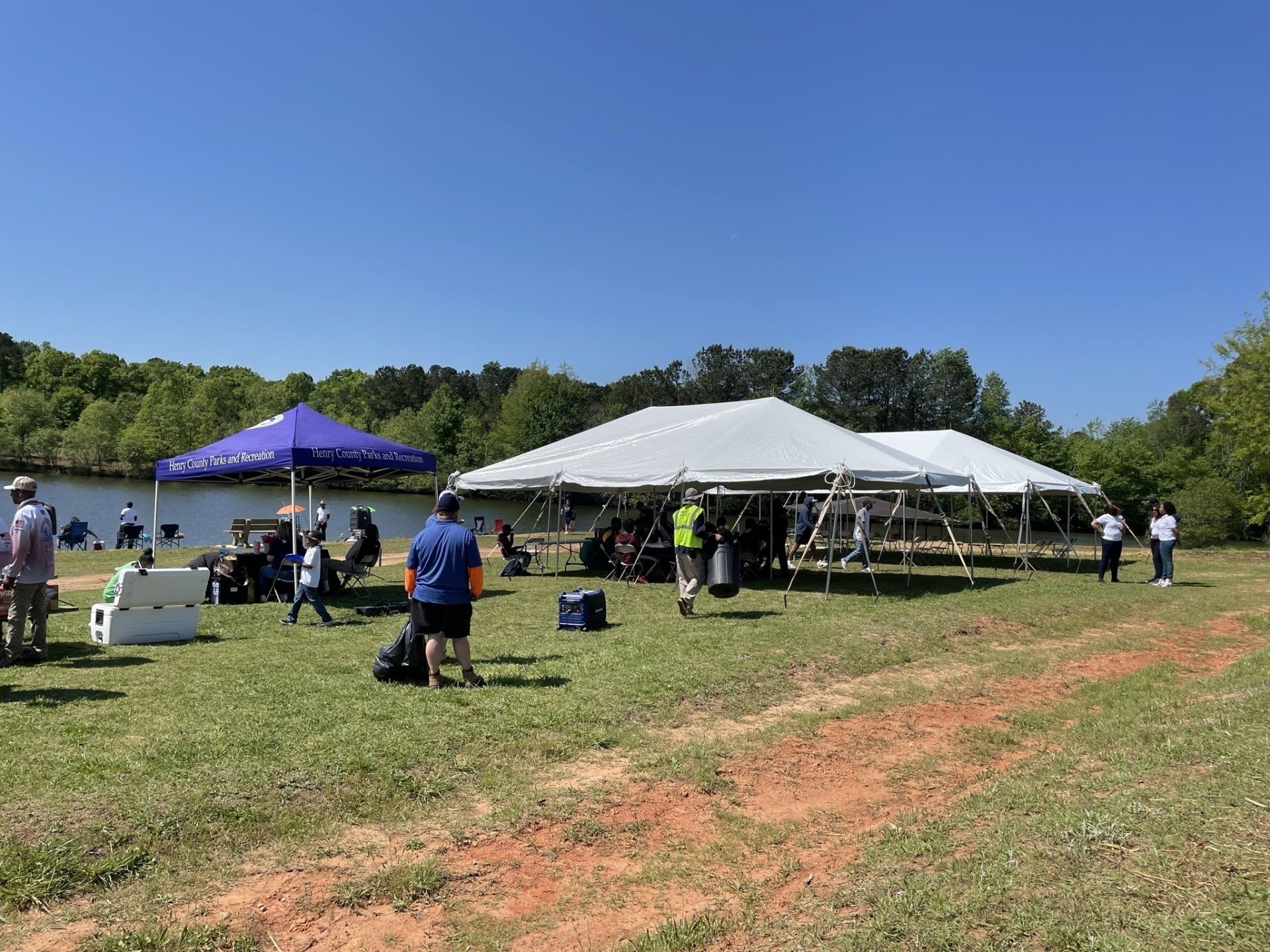 People gathered at an outdoor event under tents near a lake on a sunny day.