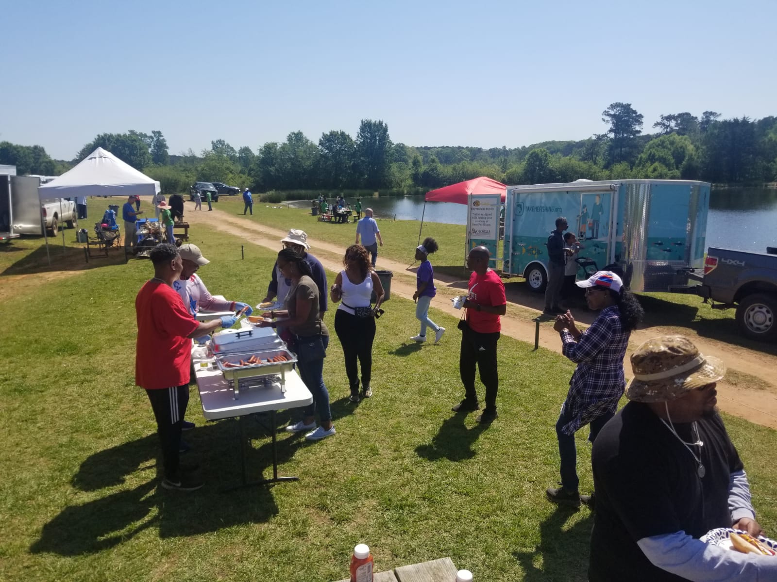 People at an outdoor gathering near a lake; food tables, food truck, sunny day.