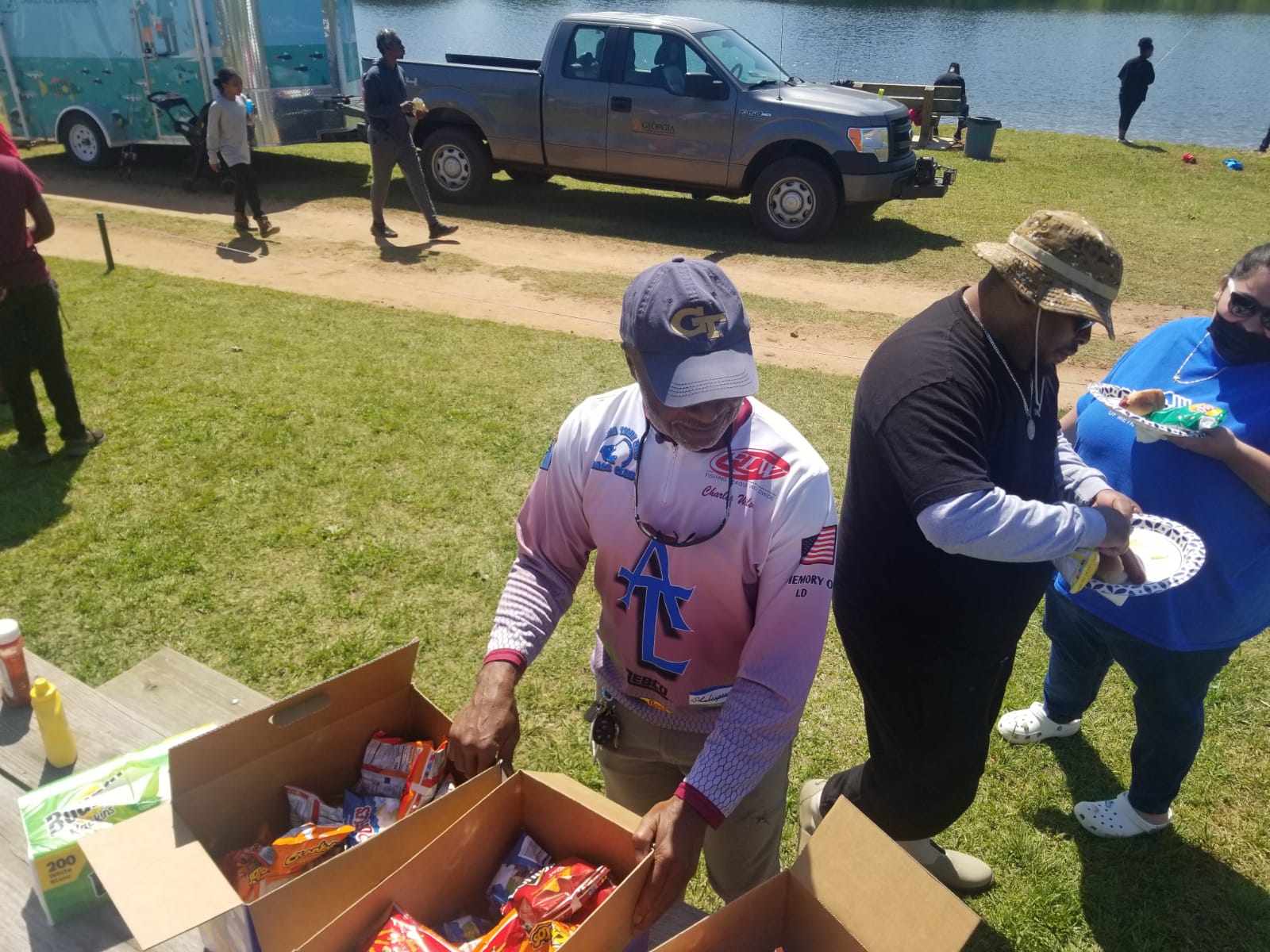 People serving food at an outdoor event near a lake. Boxes of chips and people in casual clothing.