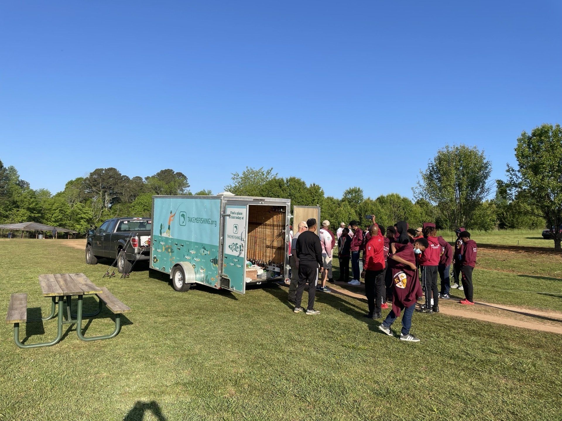 Group of students near a trailer and pickup truck on grassy area. Blue sky, trees in background.