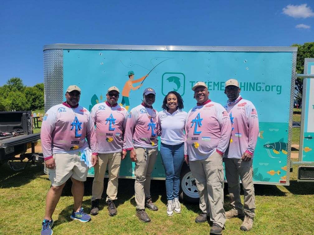 Group of people posing in front of a teal trailer with fishing logo on a sunny day.