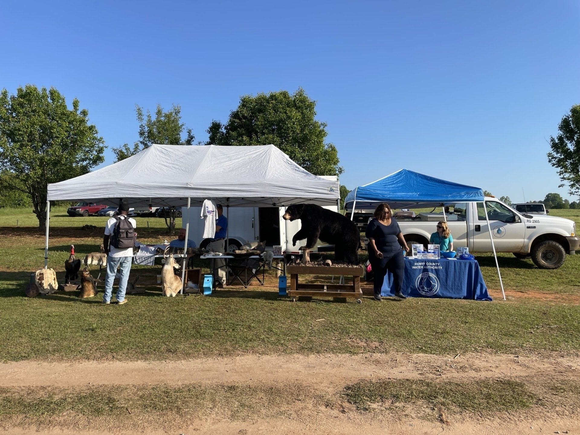 Outdoor booths at an event. People browse items under white and blue tents near a truck on a sunny day.