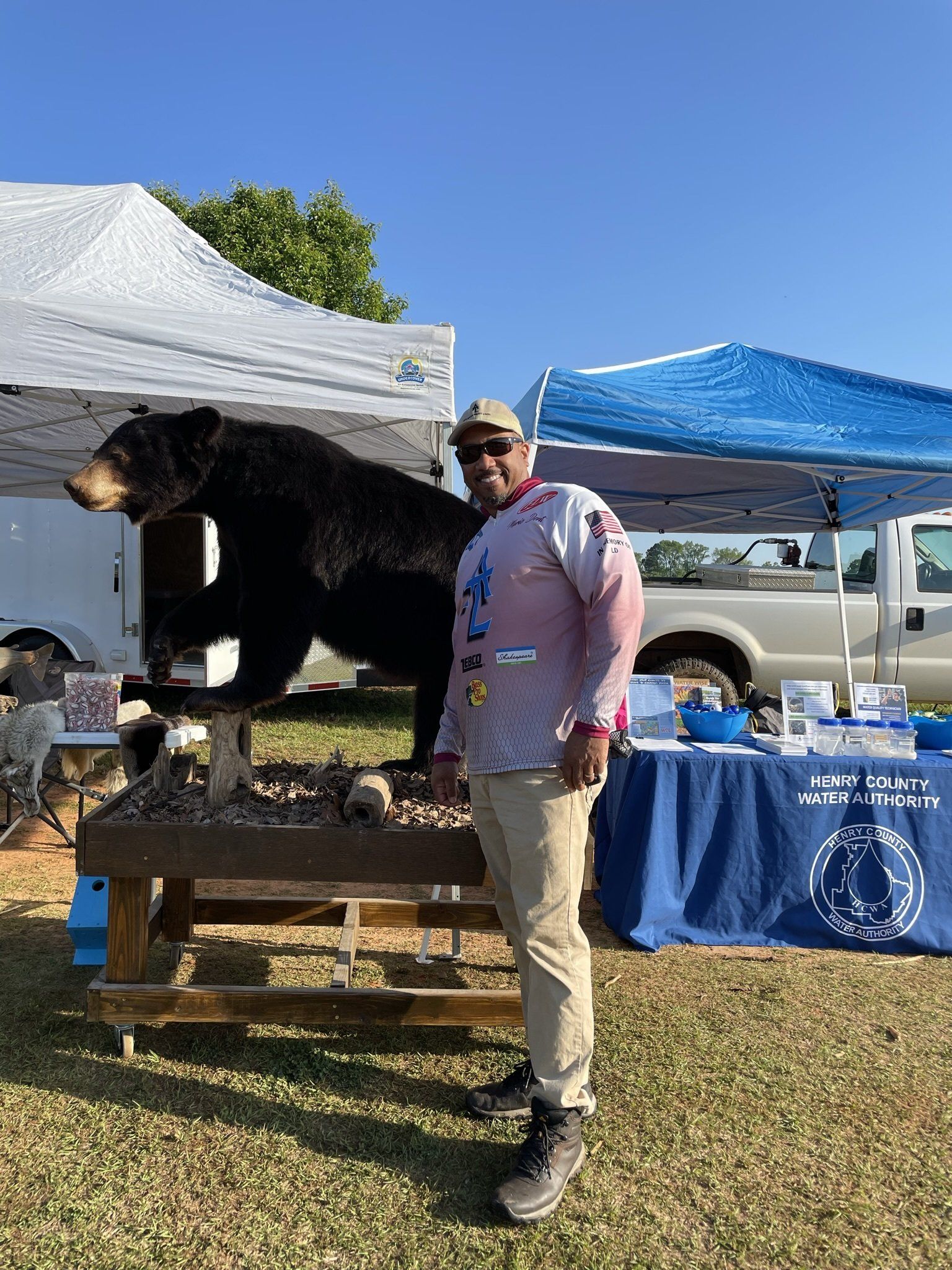 Man stands by a taxidermied black bear at an outdoor event; blue sky, white tents, tables, and information booth.
