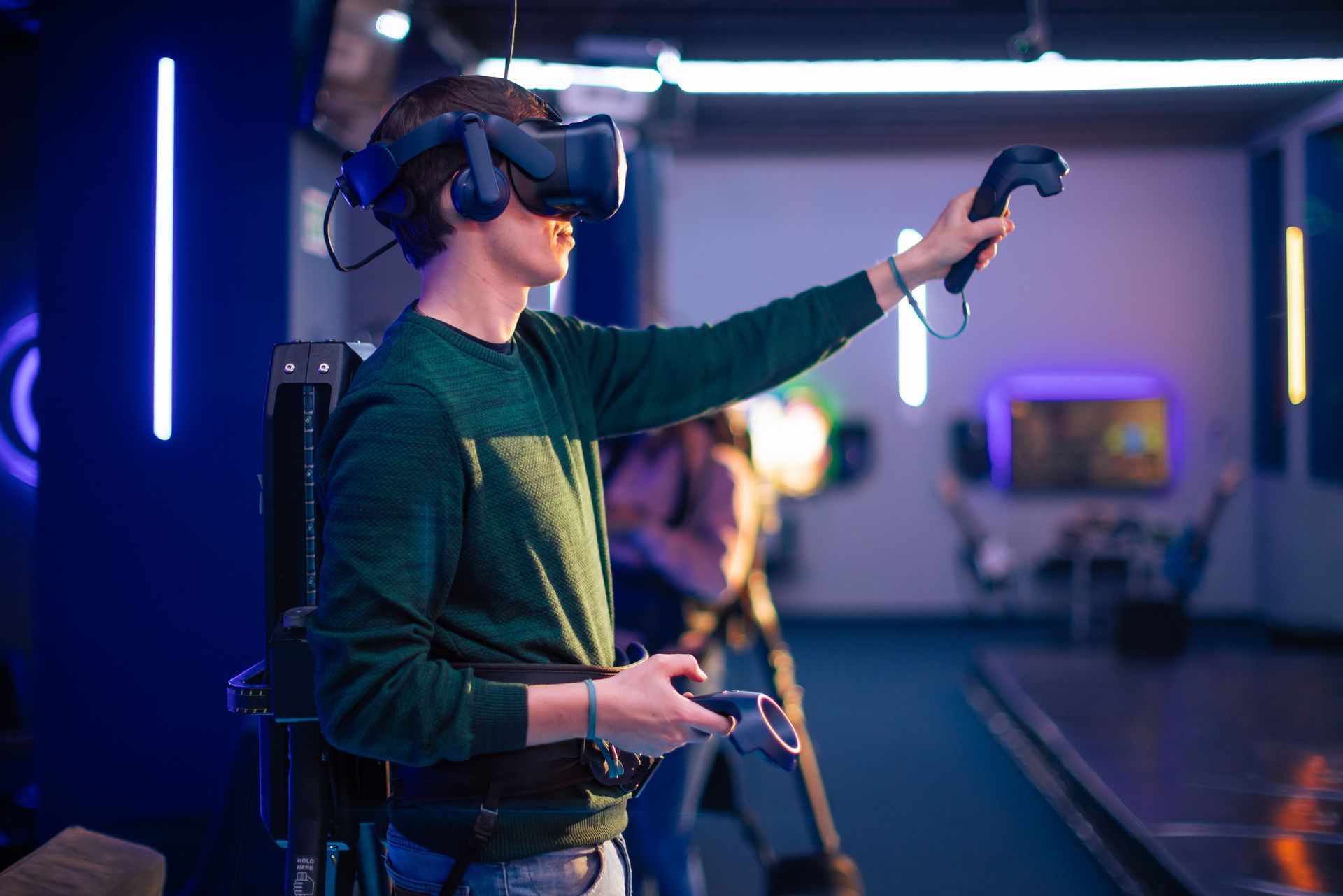 Uomo con visore VR e controller, che gioca in una stanza illuminata al neon, con un computer in stile zaino.
